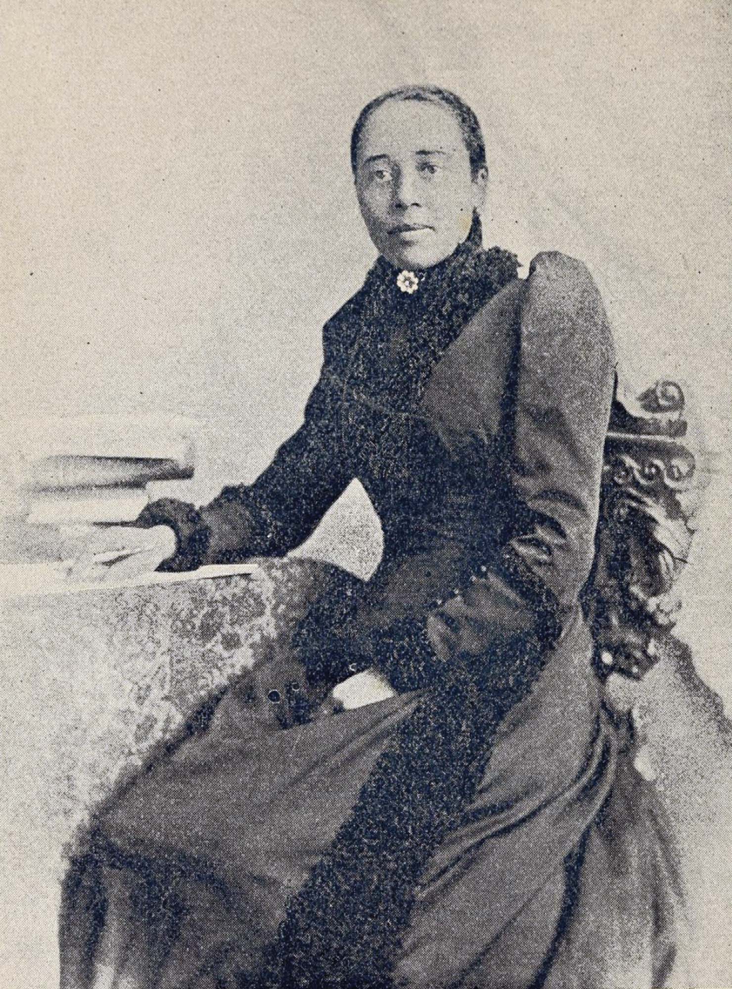 A portrait of an African American woman, wearing a long black dress. She is seated, with one arm resting on a table. Beside her is a stack of books.