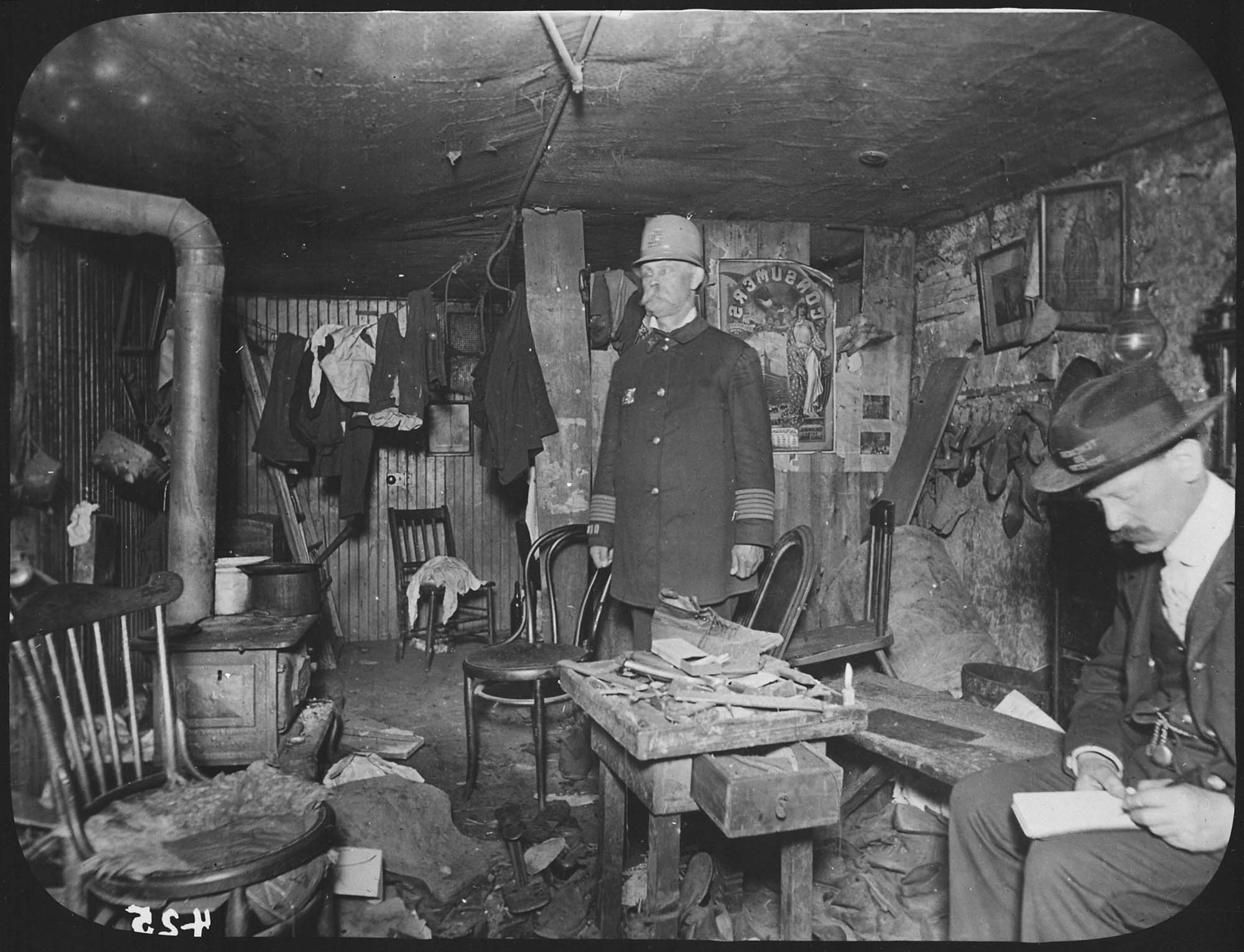 Photograph of two officials inspecting an incredibly small, dark apartment in New York City. The apartment is essentially one small room. There is a small woodstove, and clothing hanging on a line in the corner.