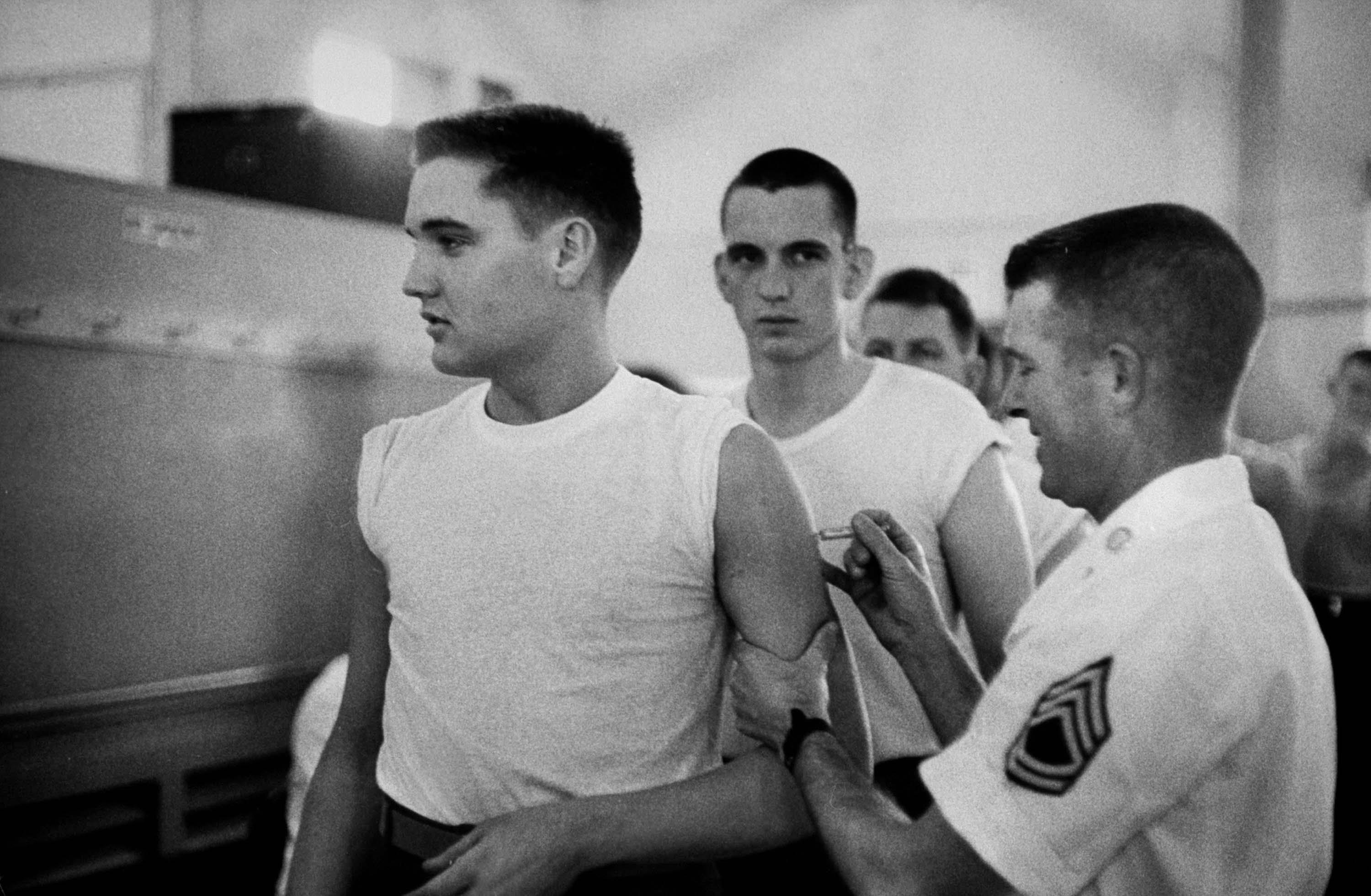 Photo of Elvis Presley, a new member of the US Army, being given an injection by a military doctor. Behind him are a line of other young men, each waiting for their turn.
