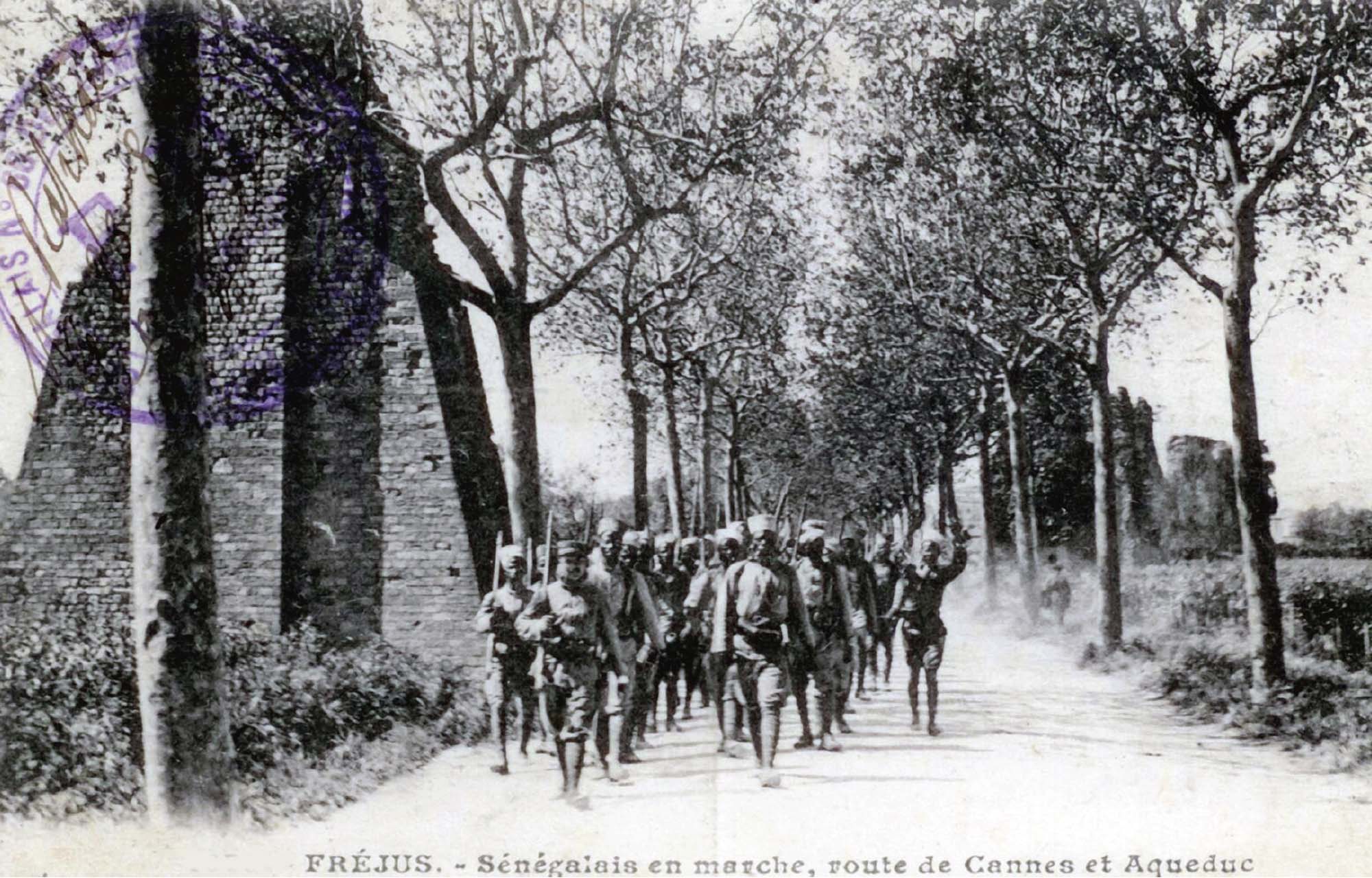A photograph of several men marching, carrying guns on their shoulders. They are walking on a tree lined road. 