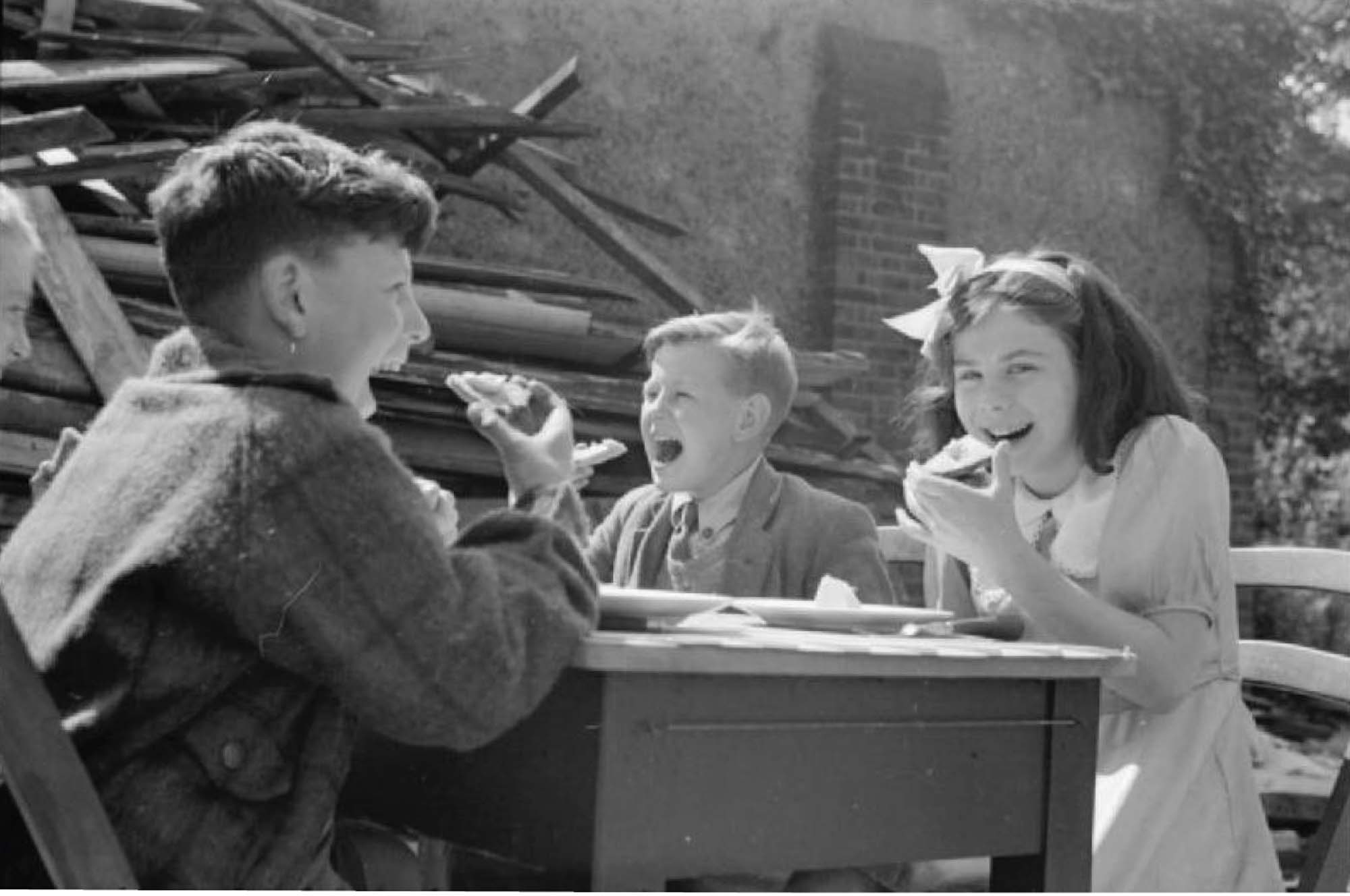 Photograph of three children, sitting at a small desk, smiling and holding their lunch as if about to take a bite. They sit outside, next to a pile of rubble.