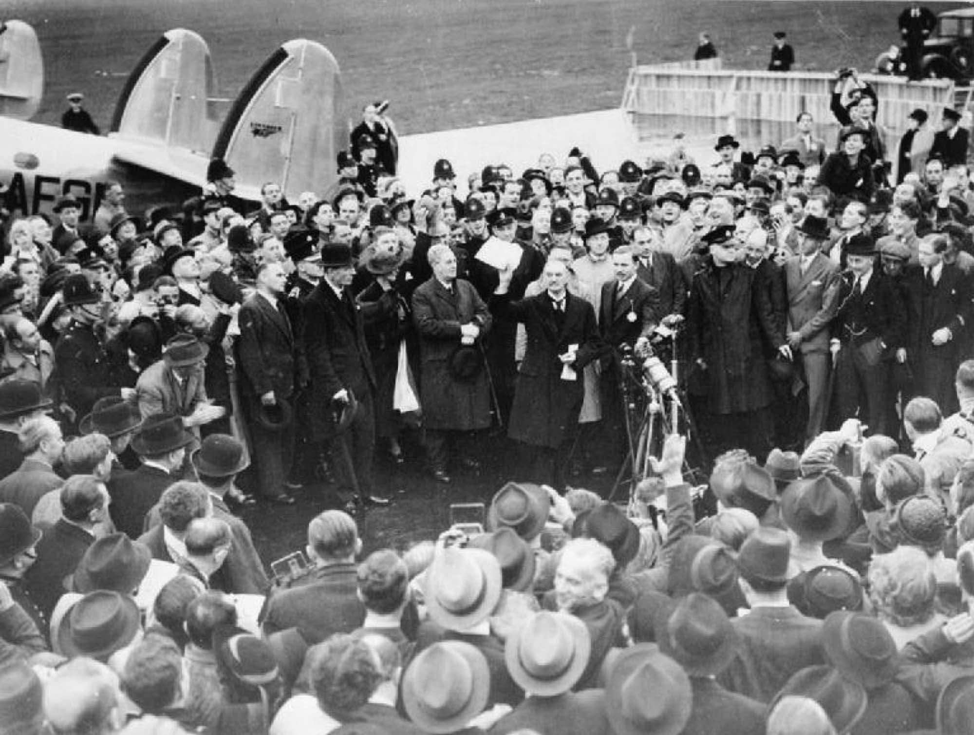 Photo of British Prime Minister Neville Chamberlin standing before a large crown, holding up the Munich Agreement.