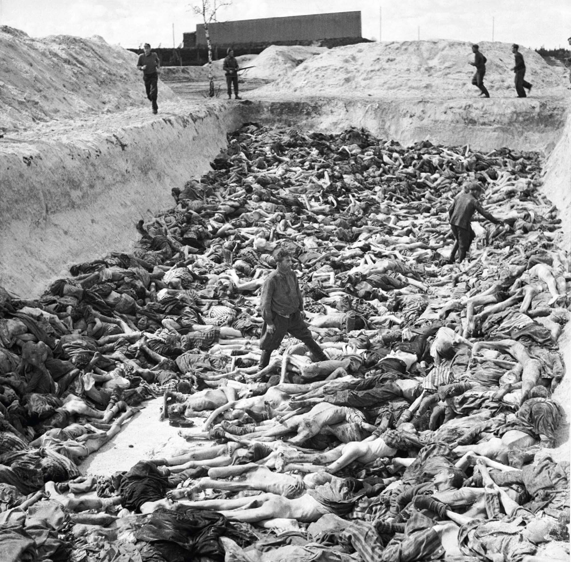 A photograph of a large trench filled with bodies of those that were murdered in one of the concentration camps. Two men walk around the mass grave.
