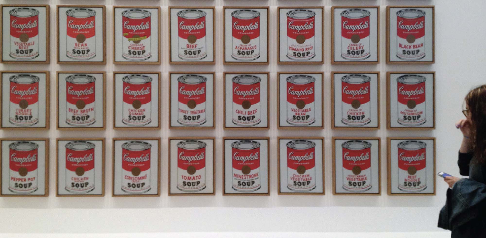 Photo of a woman standing in front of paintings of a red and white soup can in a grid formation.