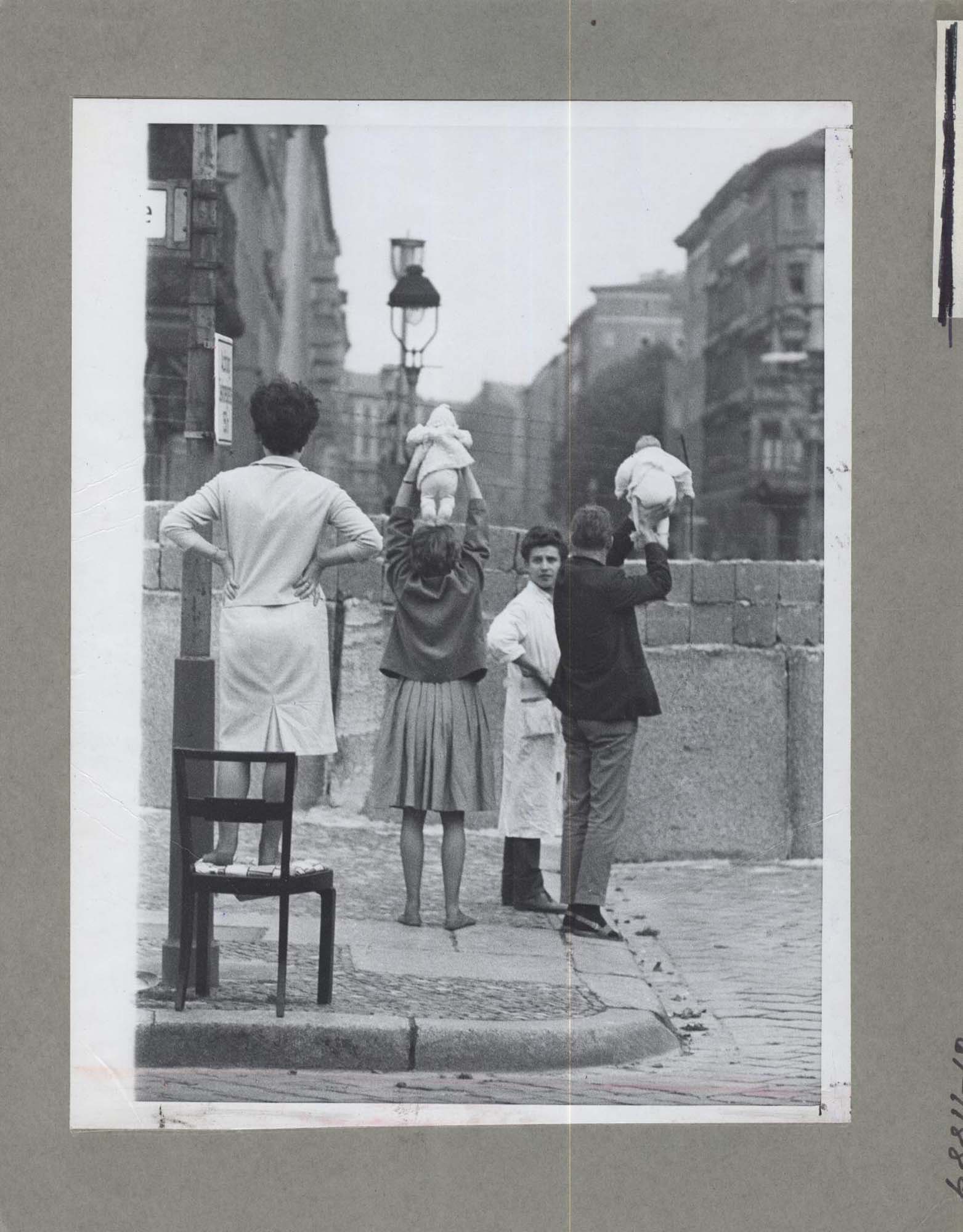 A photo of four people, standing in front of a stone wall that divided East and West Berlin. Two of the people lift their babies up above the wall.