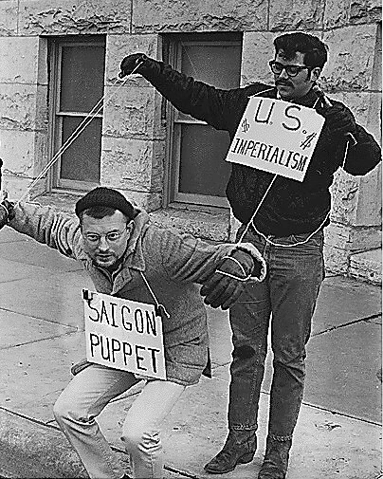 A photo of two men protesting: One man wears a sign that reads “US Imperialism” and is holding up the arms of another man, wearing a sign that reads “Saigon puppet”, with puppet strings.