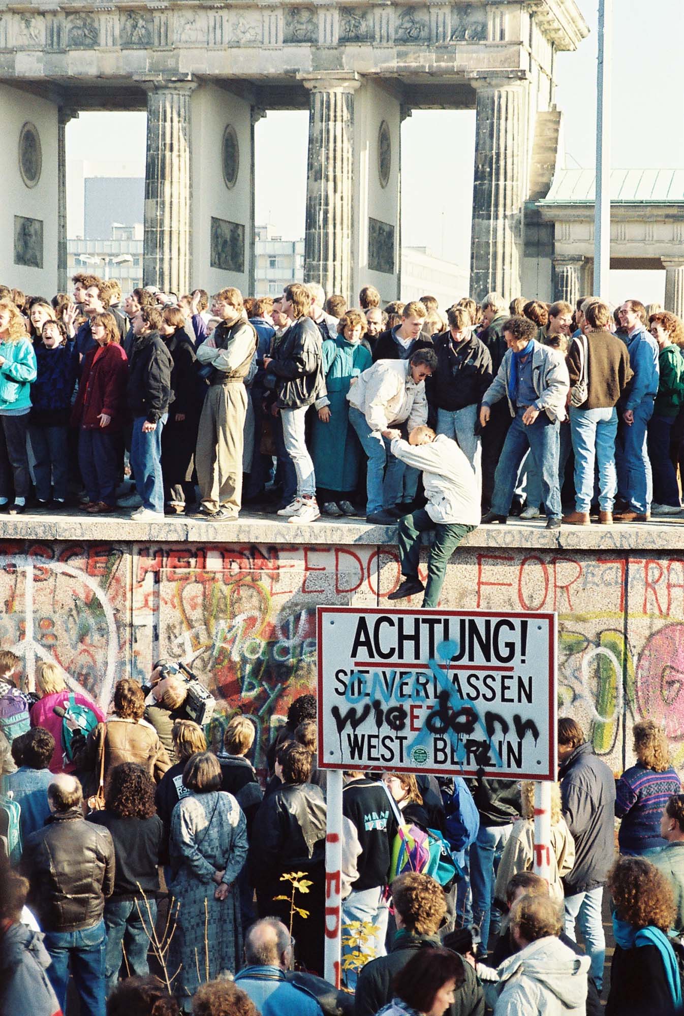 A crowd of protestors stand next to and on top of a heavily graffiti-ed wall.