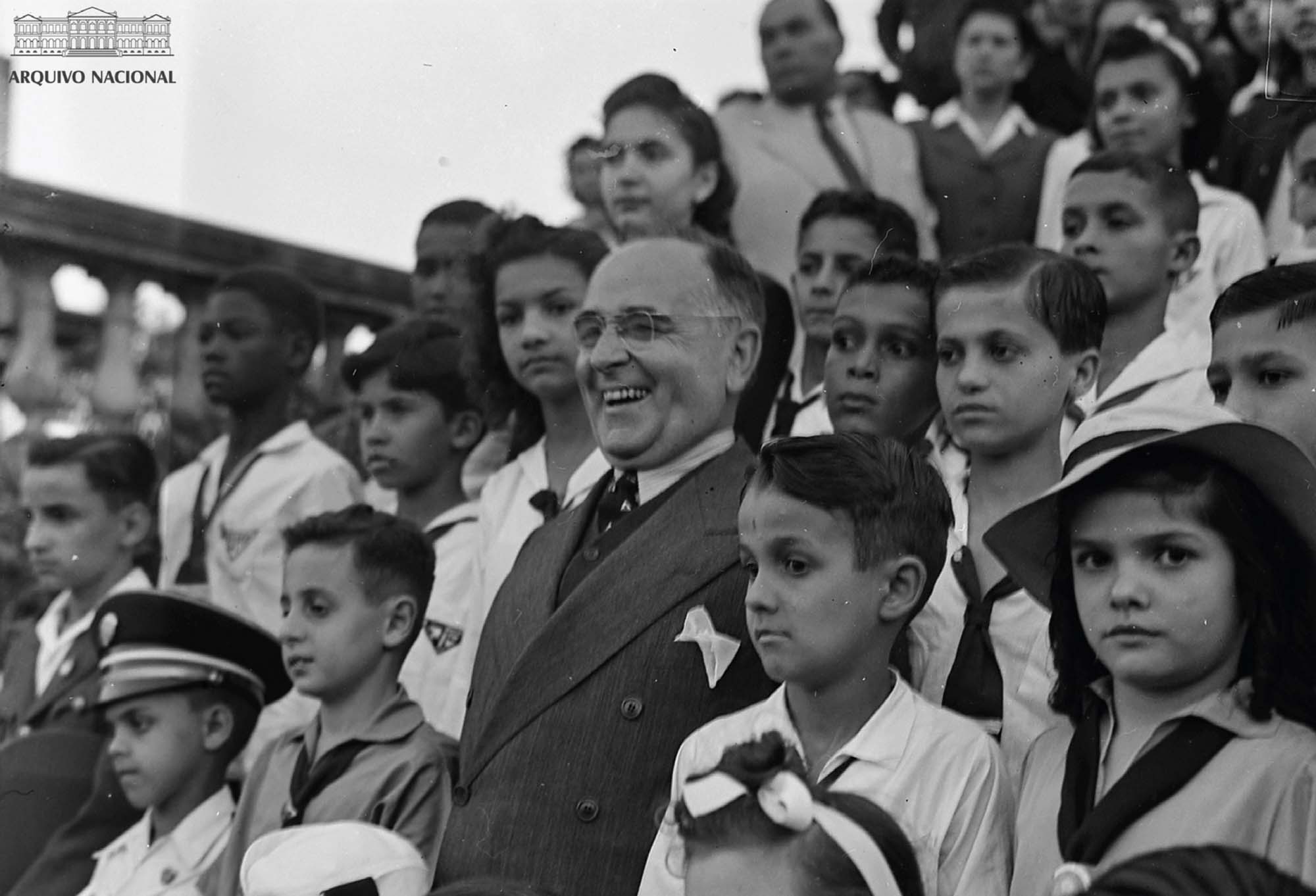 A smiling politician sanding among a crowd of children.