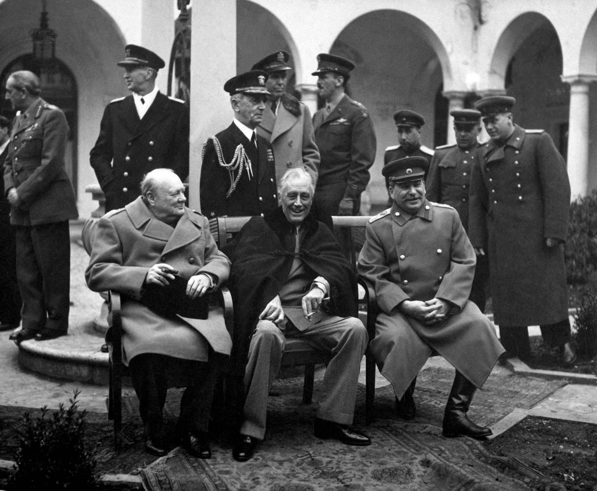 Three men, British Prime Minister Winston Churchill, US President Franklin Roosevelt, and Soviet leader Joseph Stalin seated in chairs while high ranking military officers stand behind them.