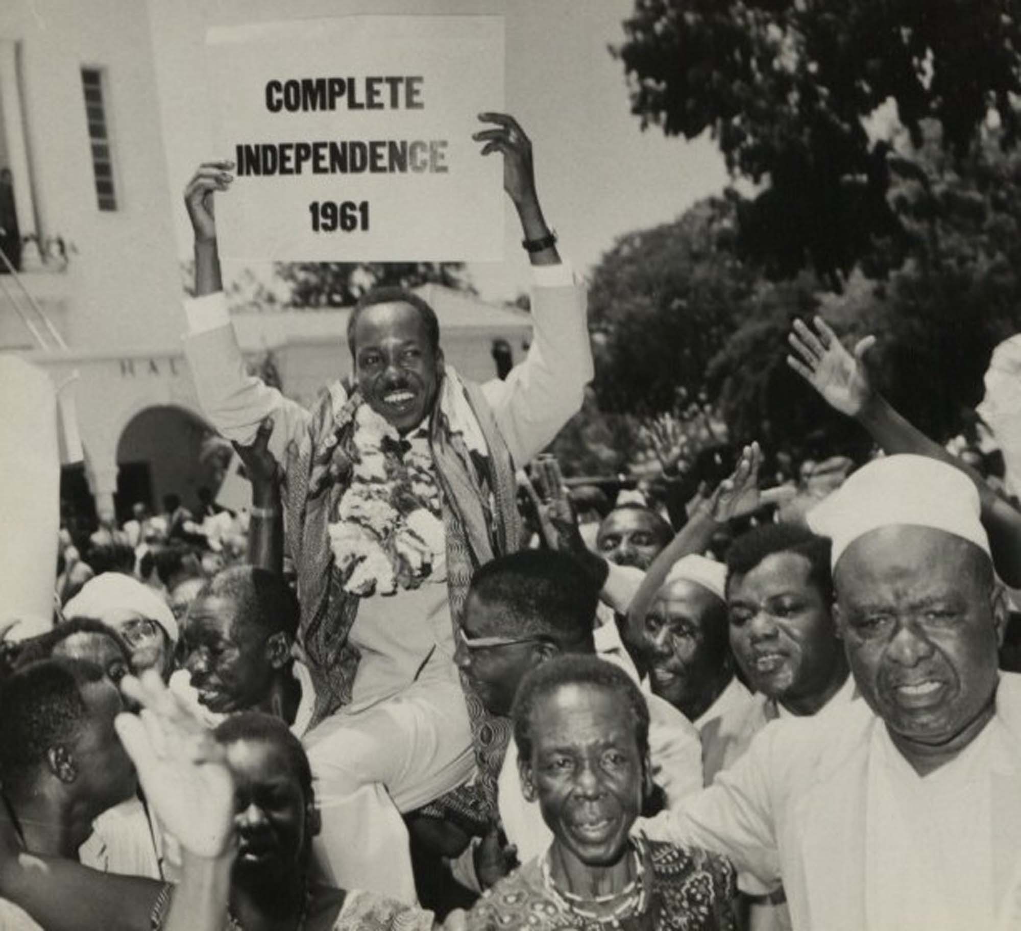 A jubilant crowd holds a man on their shoulders. The man carries a sign celebrating “Complete Independence 1961.”