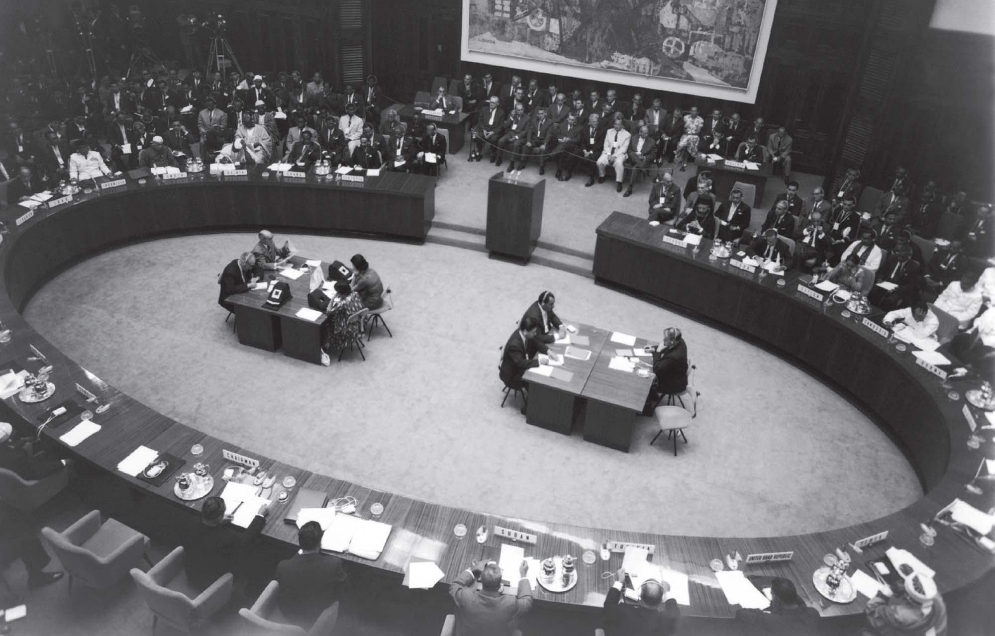 Overhead view of a large, oval conference table with delegates sitting around in discussion.