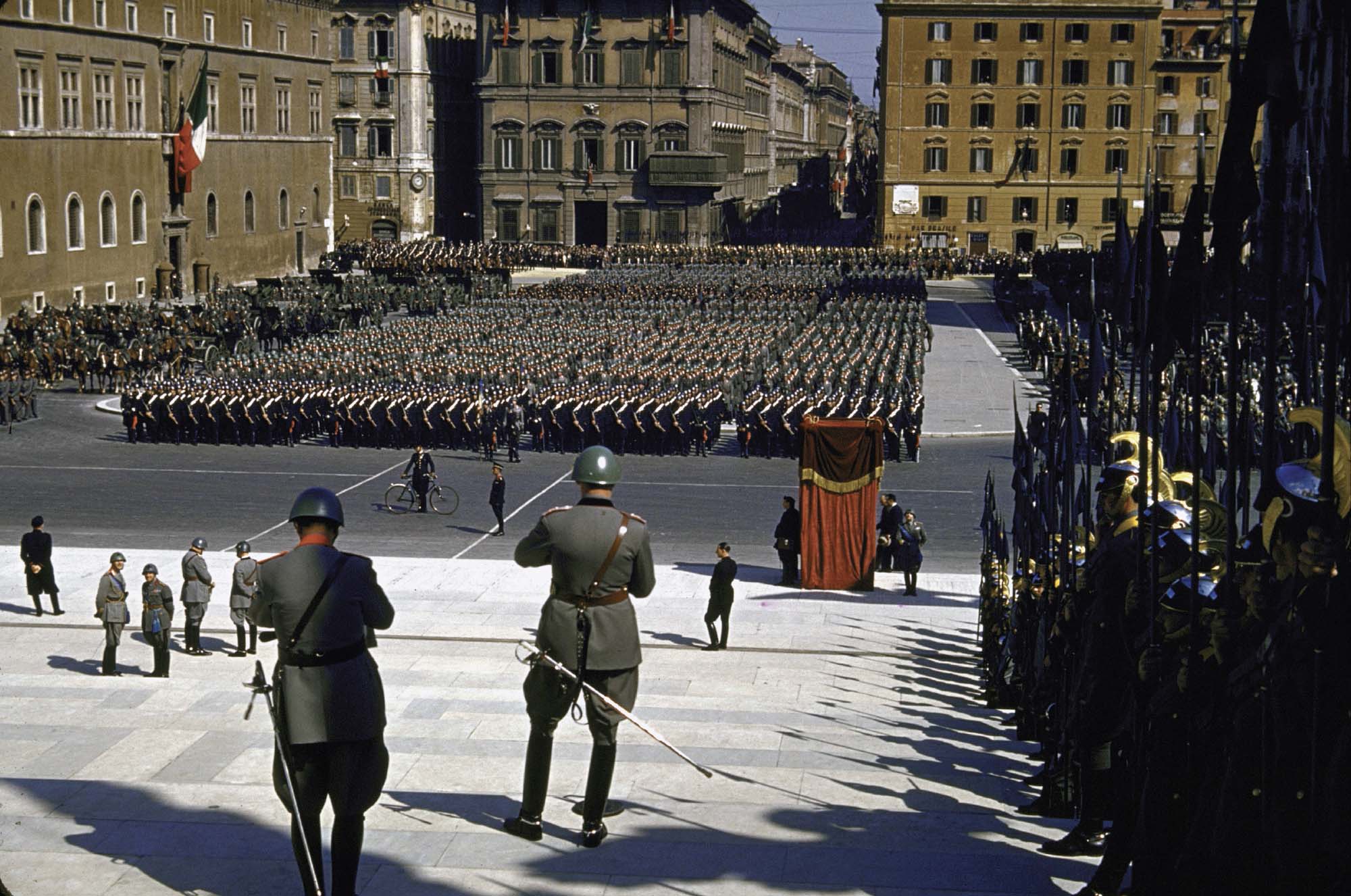 A photograph of a large, organized parade in the center of a town. Several men in uniforms and helmets stand watching the parade.