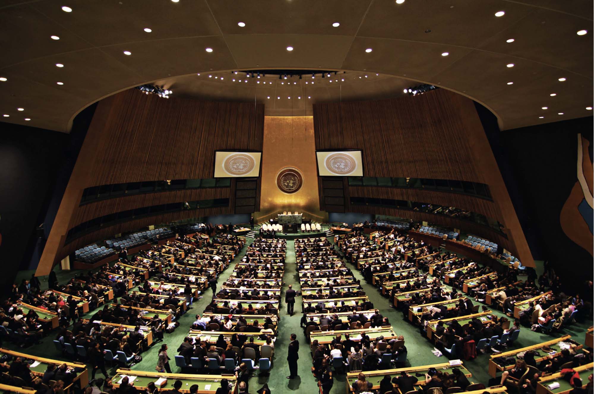 A photograph shows a large auditorium, with rows of seats. There is green carpeting and several spotlights on the ceiling. All of the seats are filled, and all are facing a podium at the back of the auditorium.