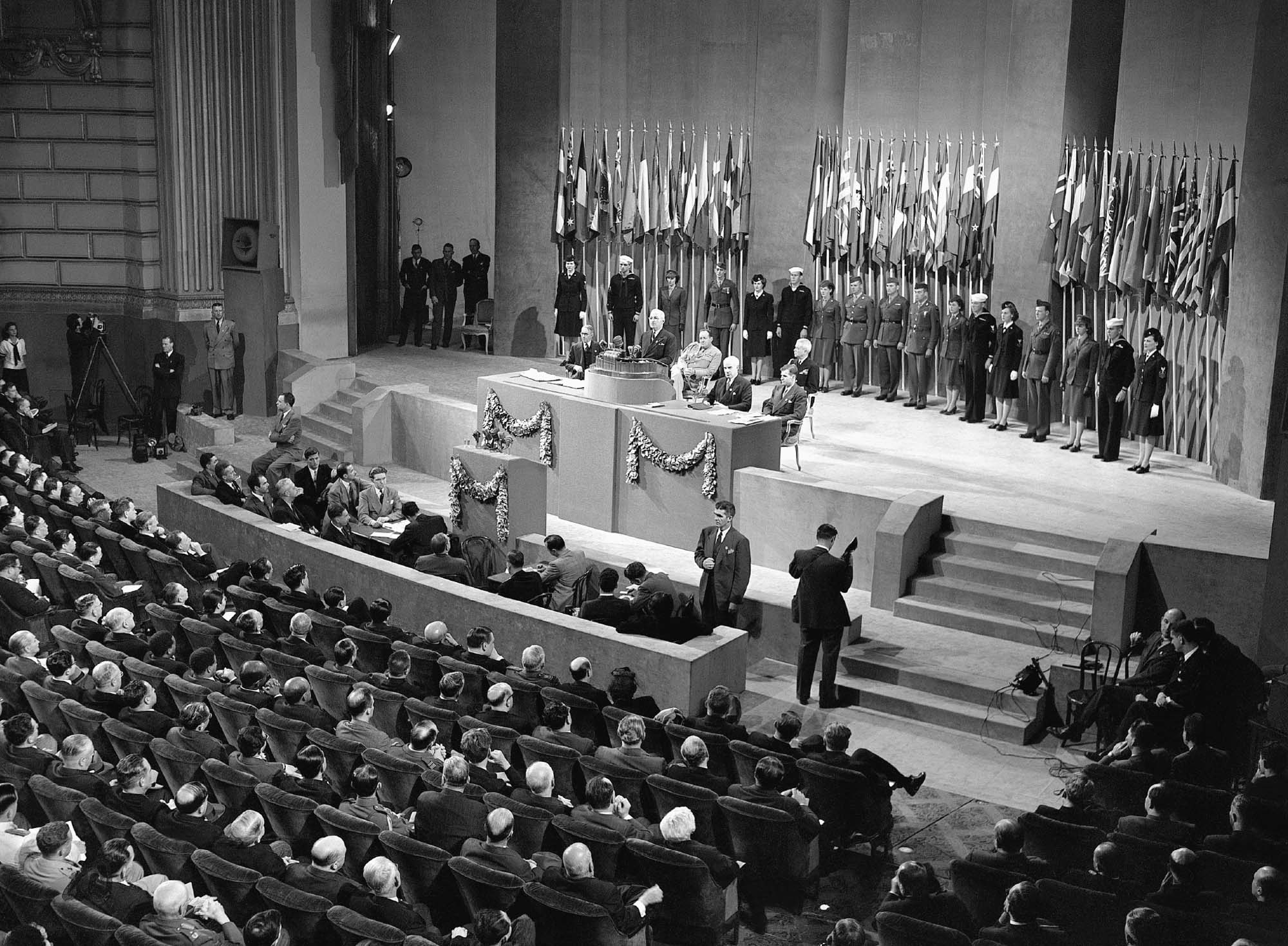 Inside an auditorium, a man stands at a podium on a stage, with several others seated next to him. Behind them, on the stage, are several others standing in front of flags representing the United Nations. The auditorium seats are filled with people watching the presenters.