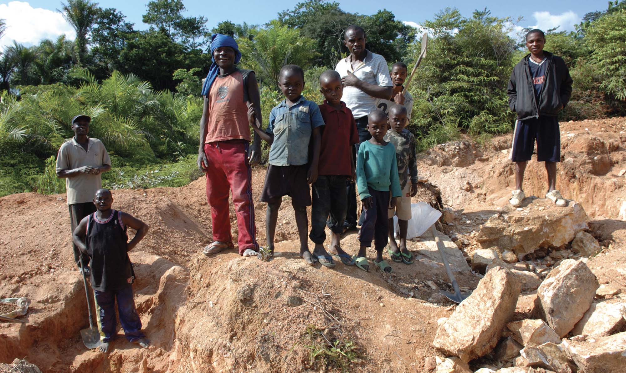 A photo of several boys and men standing on red-colored dirt and rocks. They are looking at the camera, and behind them is lush greenery.