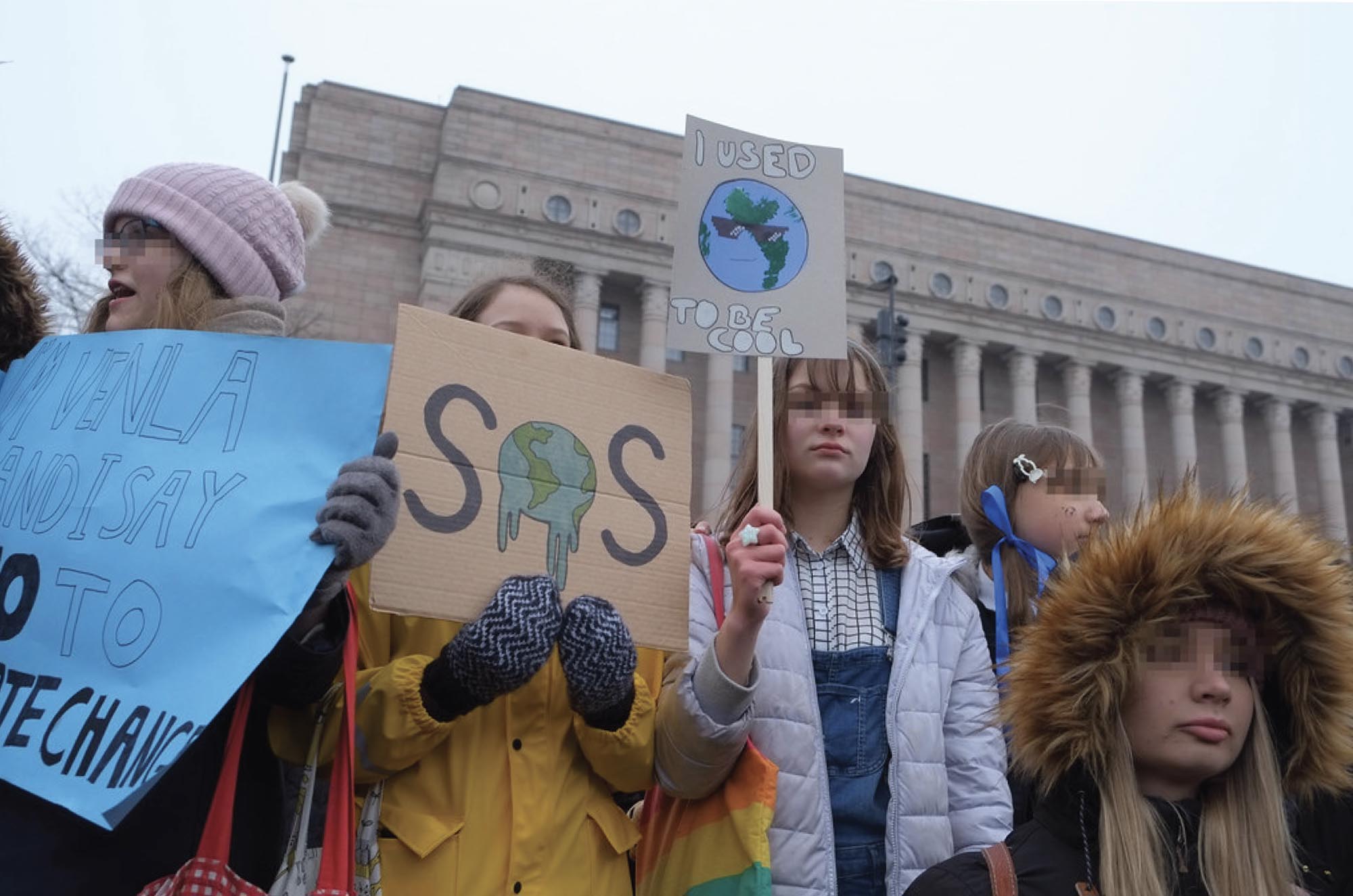 A photograph of a group of young girls protesting at a climate strike, holding up handmade signs. One sign reads “SOS”, the ‘O’ replaced by a melting planet Earth.