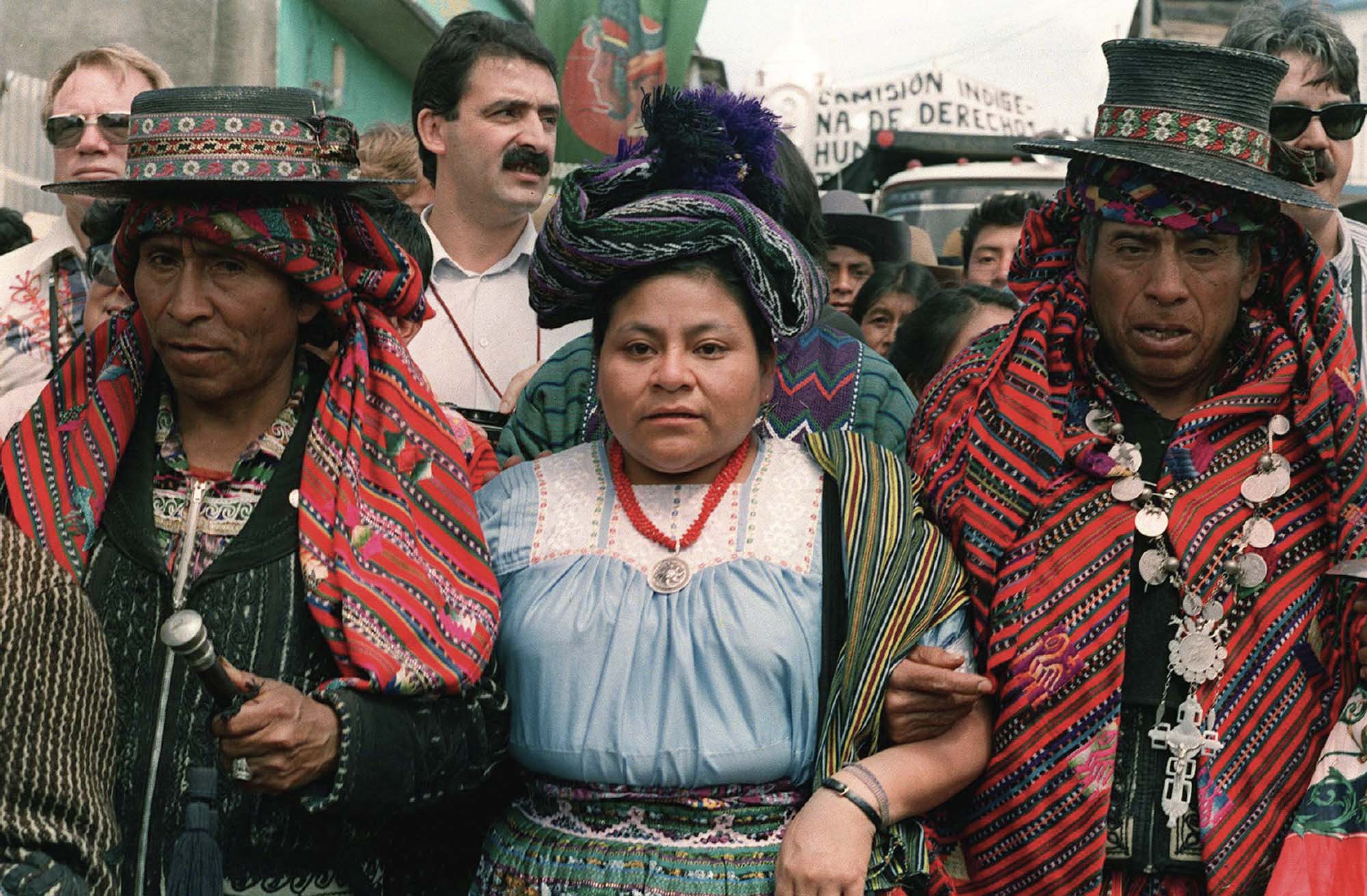 Photo of a crowd of people, featuring two men and one woman wearing ethnically specific Guatemalan clothing.