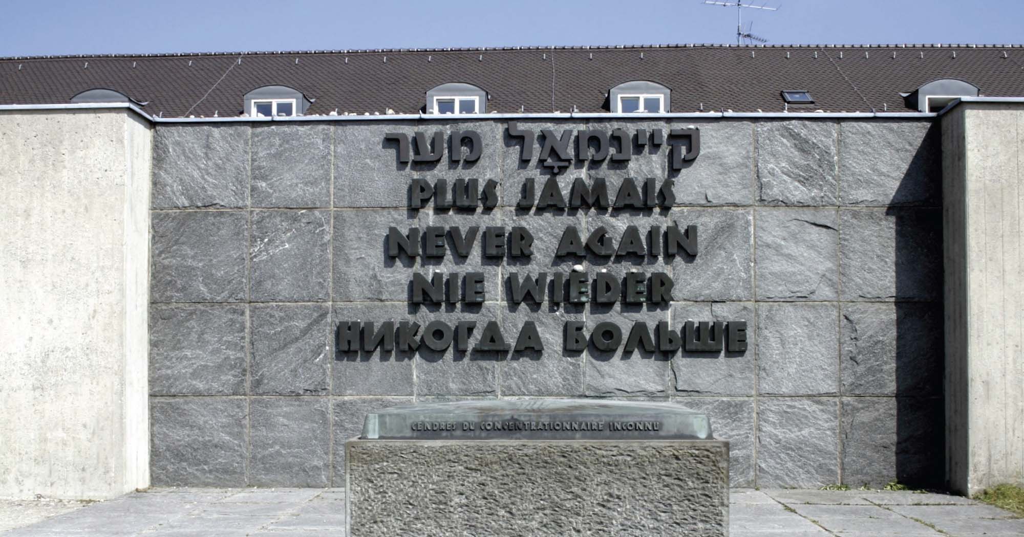 A photograph of a stone memorial. It is built of several large stone blocks and reads “Never again” in several languages.
