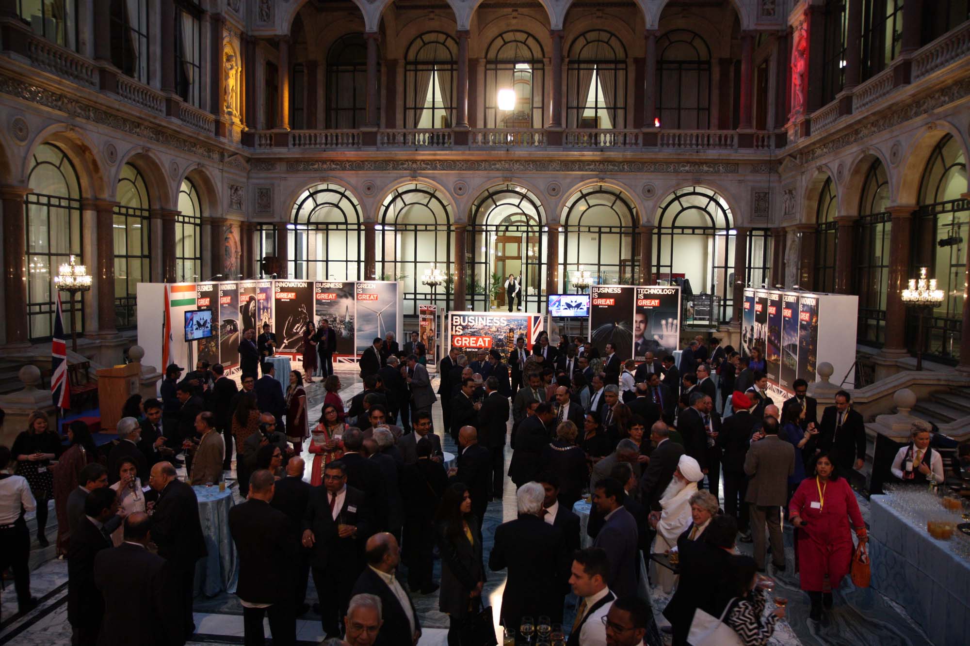 Photo of an Indian Diaspora event from 2014 in which a large group of Indian people are shown socializing at the London Foreign and Commonwealth Office. Large posters promoting business, innovation, and technology can be seen in the background.