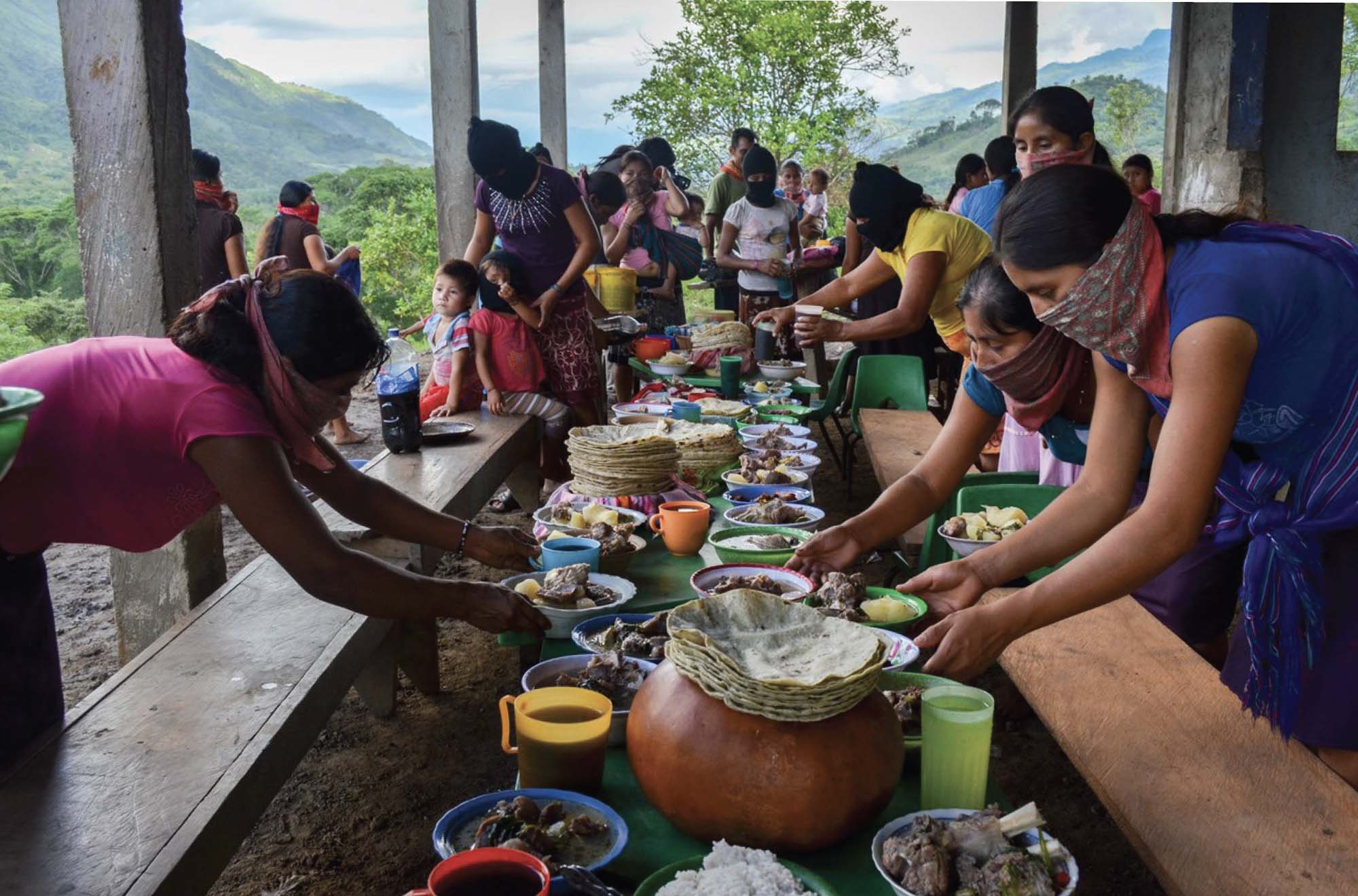 Several people set a table under a wooden shelter. Most are wearing handkerchiefs around their noses and mouths  and are filling the table with a large feast. Behind them is a view of a lush, mountainous area.