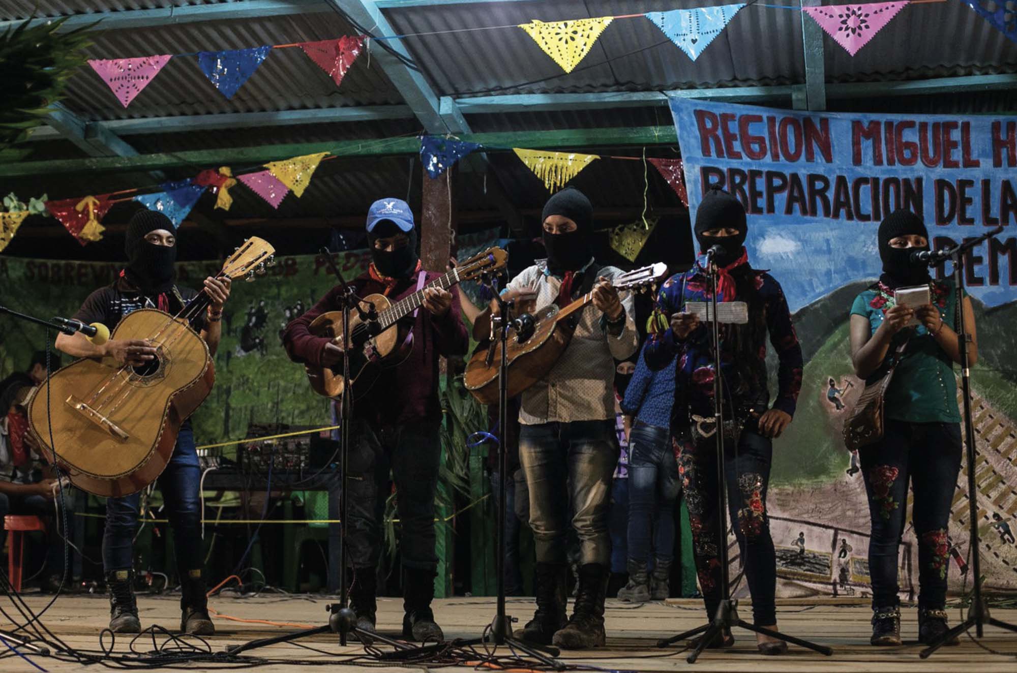 Five musicians, wearing coverings over their noses and mouths, stand on a stage in front of microphones. Three are playing guitars of various sizes and two are holding notepads.