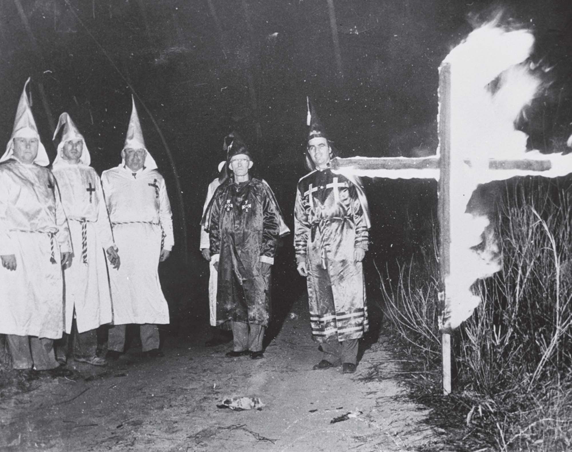 Five figures wearing hoods and robes of the KKK standing in front of a burning cross.