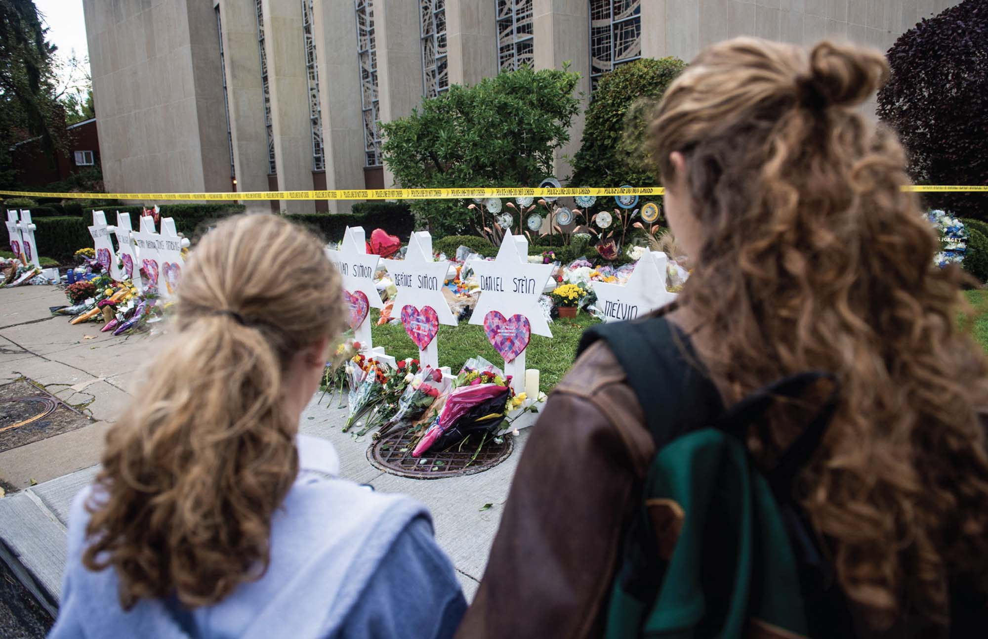 Two girls stand in front of a memorial with flowers and names of the victims of the Tree of Life Synagogue massacre.