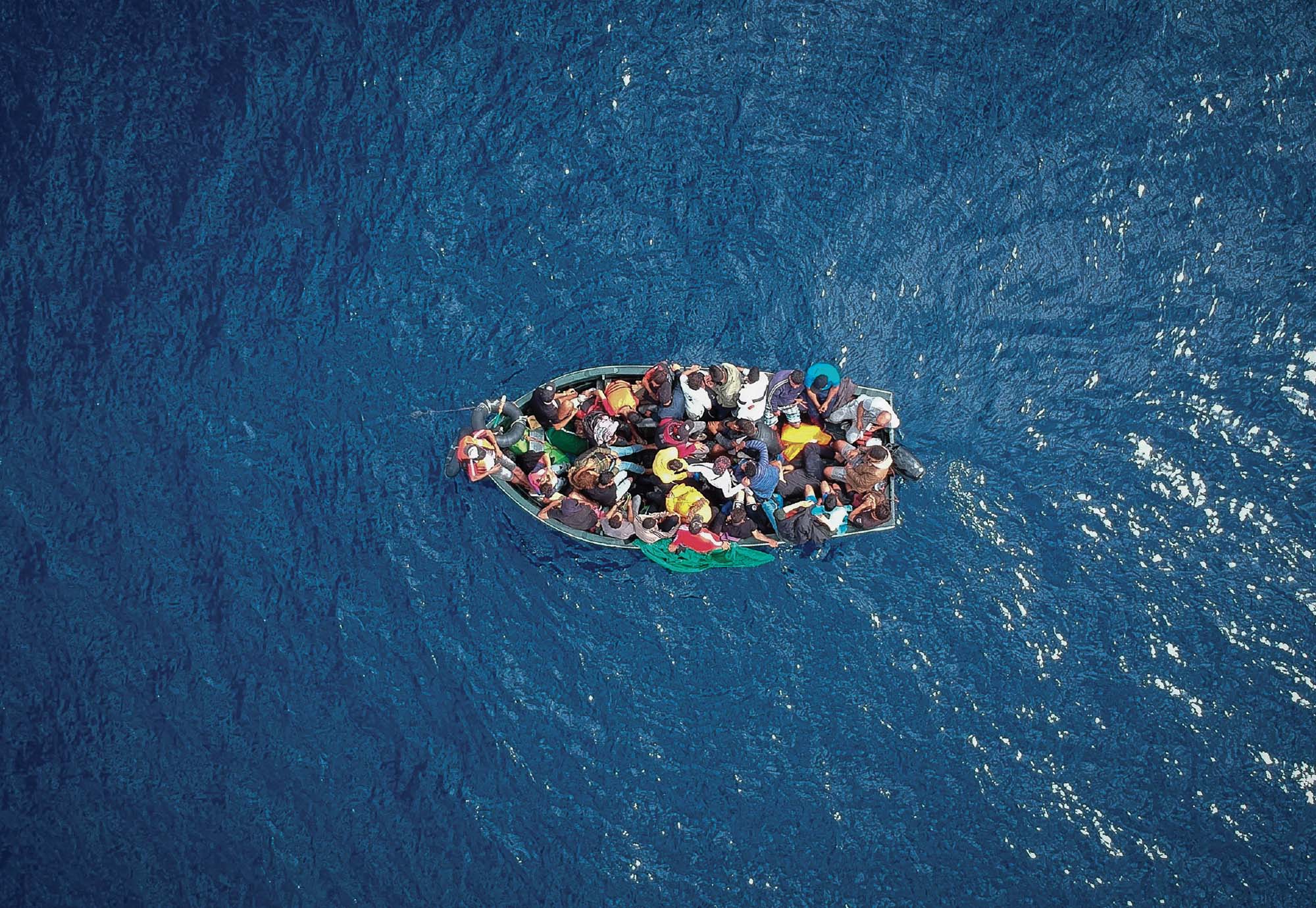 Photo of an overcrowded boat holding dozens of people in the middle of a blue body of water.