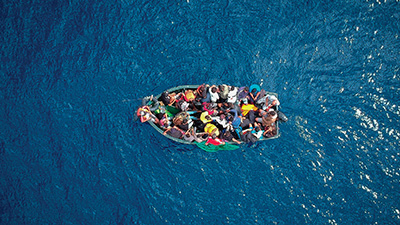 Photo of an overcrowded boat holding dozens of people in the middle of a blue body of water.