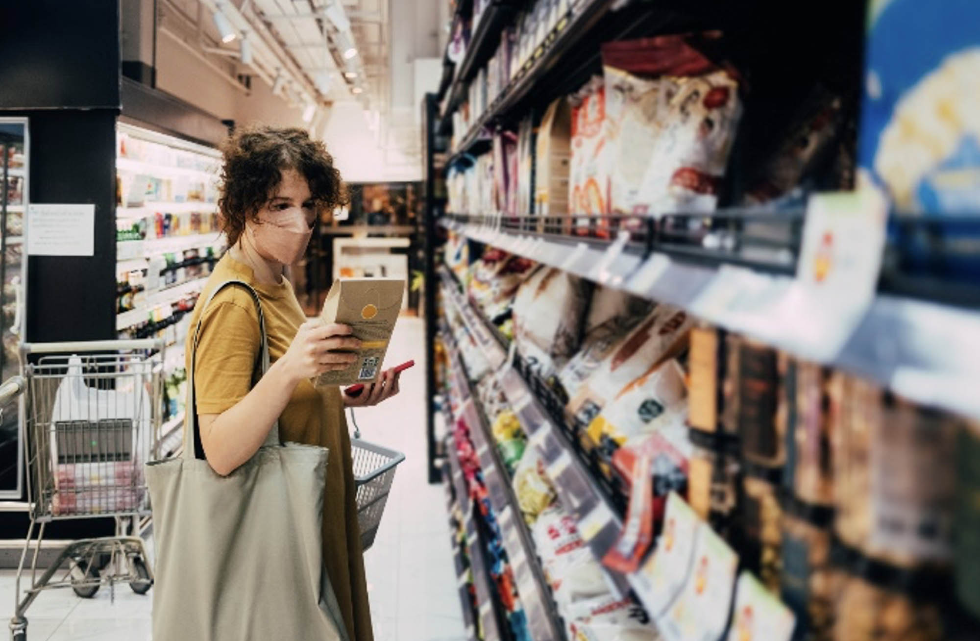 Photo of a person standing in an aisle of a grocery store, wearing a face mask and reading the label of a product.