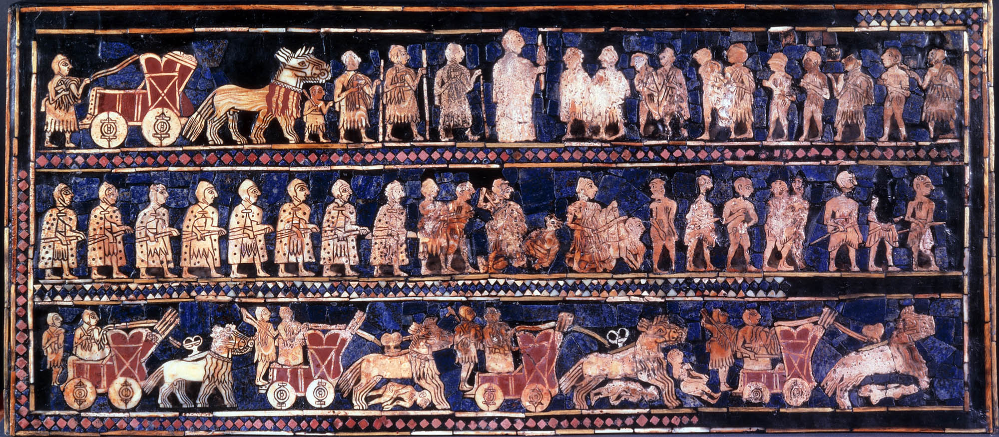 A hollow, long wooden box with inlaid with a mosaic of shell, red limestone, and lapis lazuli. This side of the box displays the "War" panel showing the king in the middle standing in front of a row of prisoners, wagons carrying a driver and a warrior.