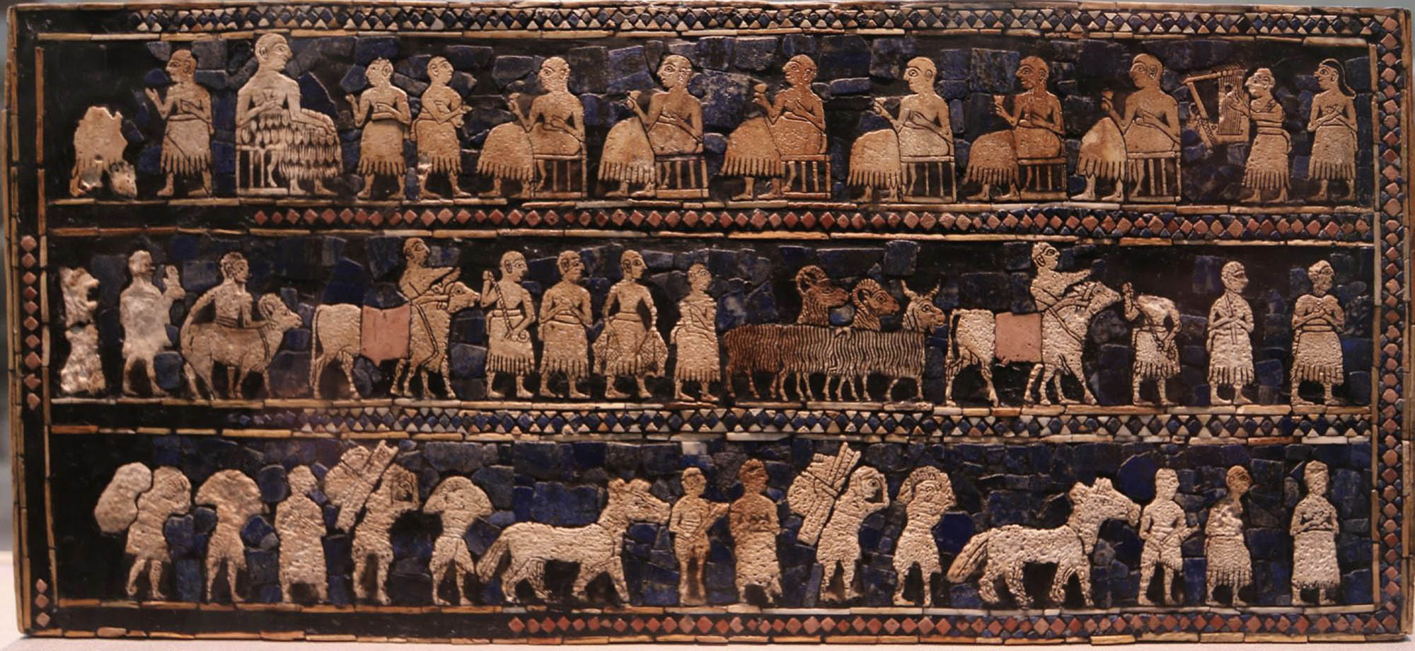 A hollow, long wooden box with inlaid with a mosaic of shell, red limestone, and lapis lazuli. This side of the box displays the "War" panel showing the king in the middle standing in front of a row of prisoners, wagons carrying a driver and a warrior.