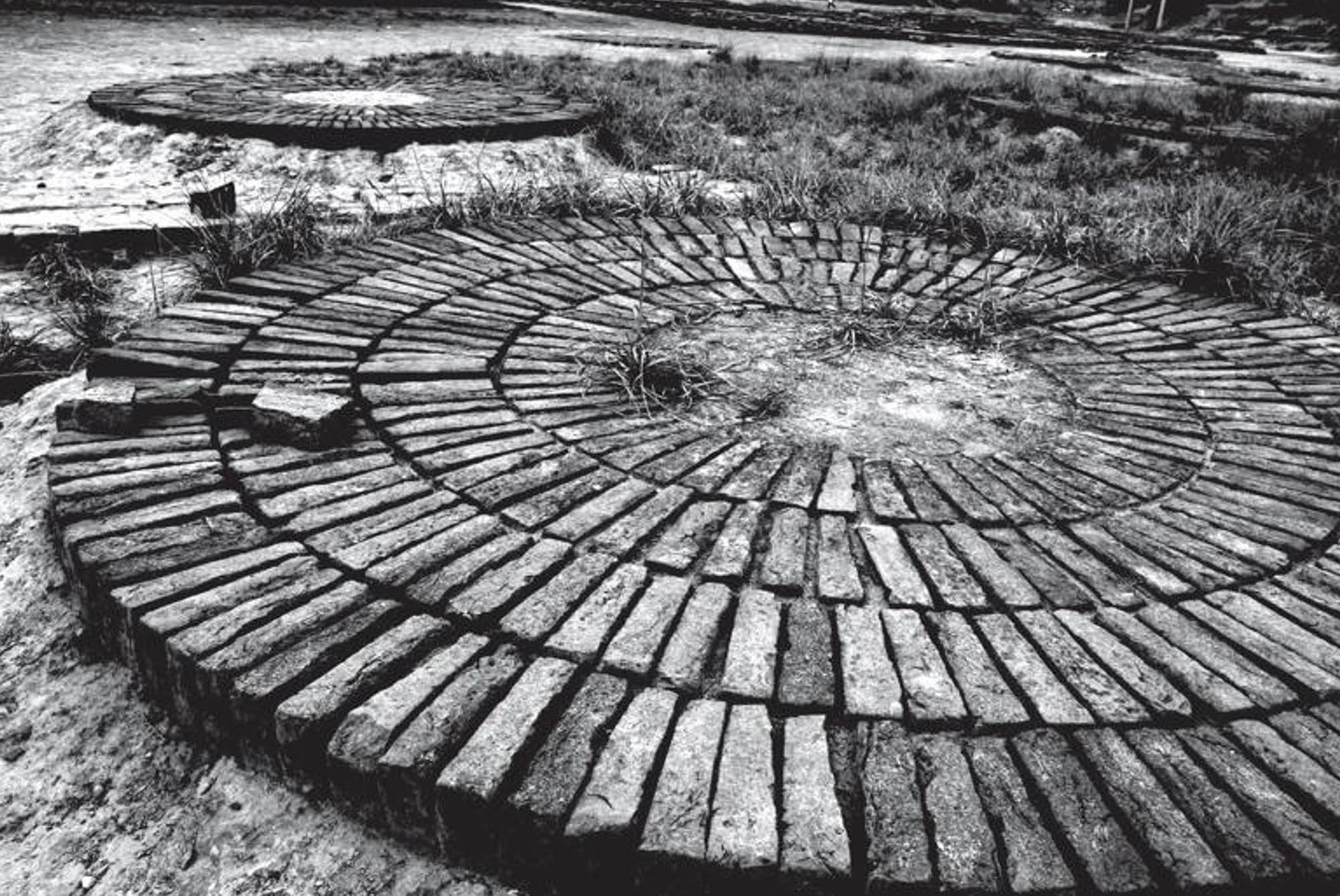 Photo shows circular arrangements of bricks surrounding a round patch of ground. Grasses are growing outside of the brick arrangements.