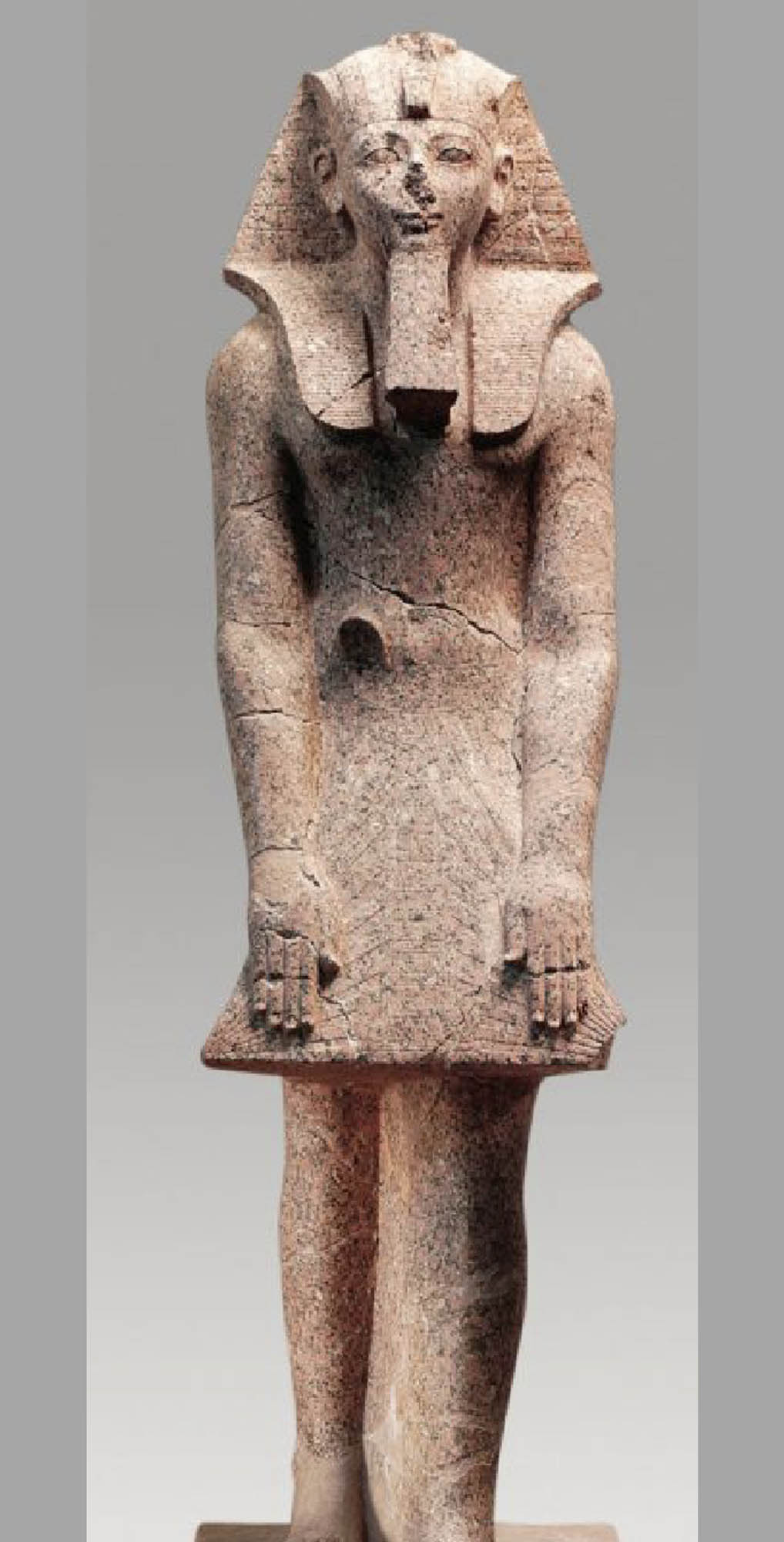 Statue of a pharaoh standing with her arms at her side, wearing a headdress and skirt. The statue has some cracks in it and her nose is broken off.