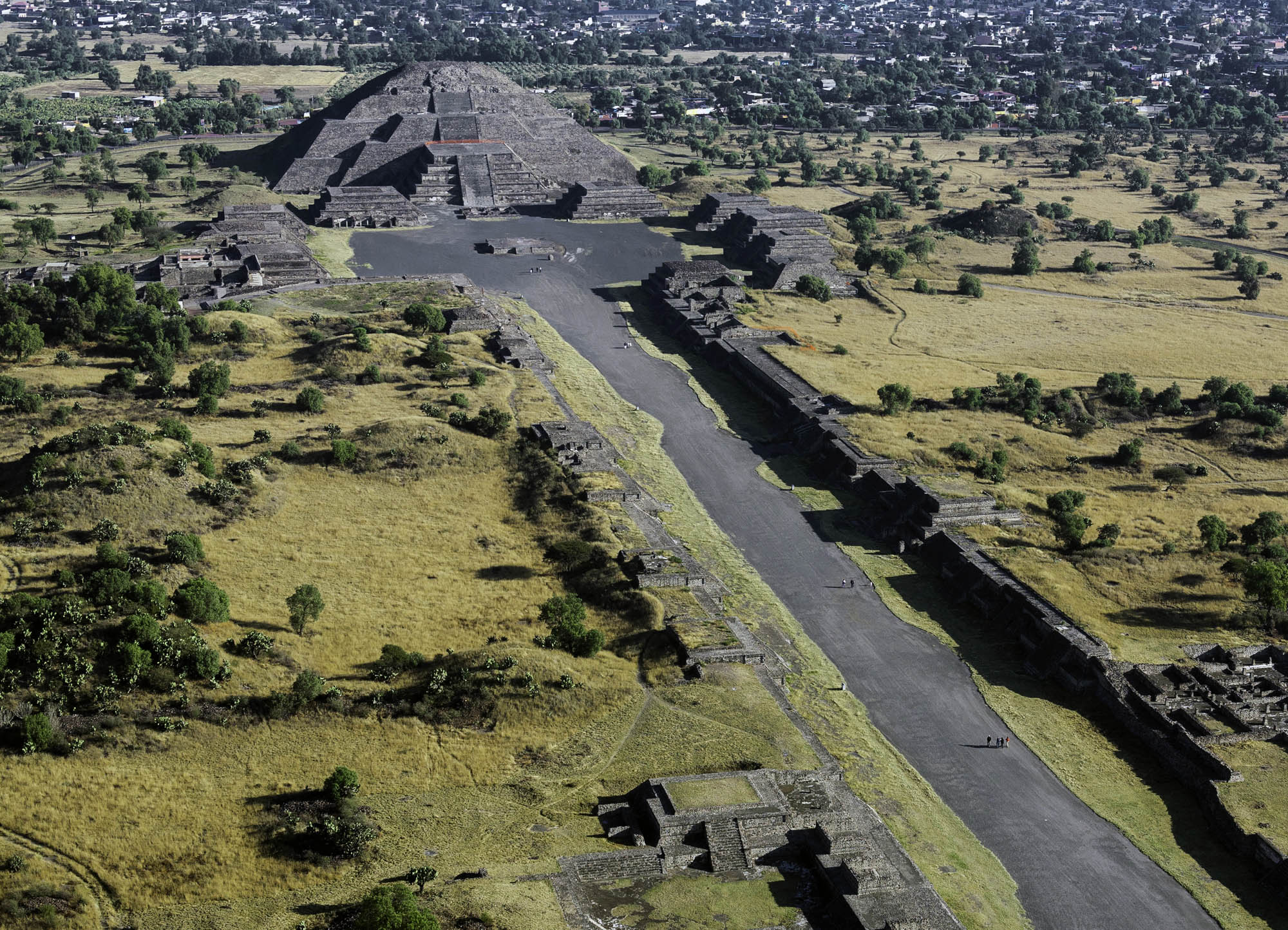 Photo of Teotihuacan, showing a long narrow road to a center area surrounded by the structures that make up Teotihuacan. There is a larger, pyramid structure, and several smaller structures that are leveled and have staircases going up them