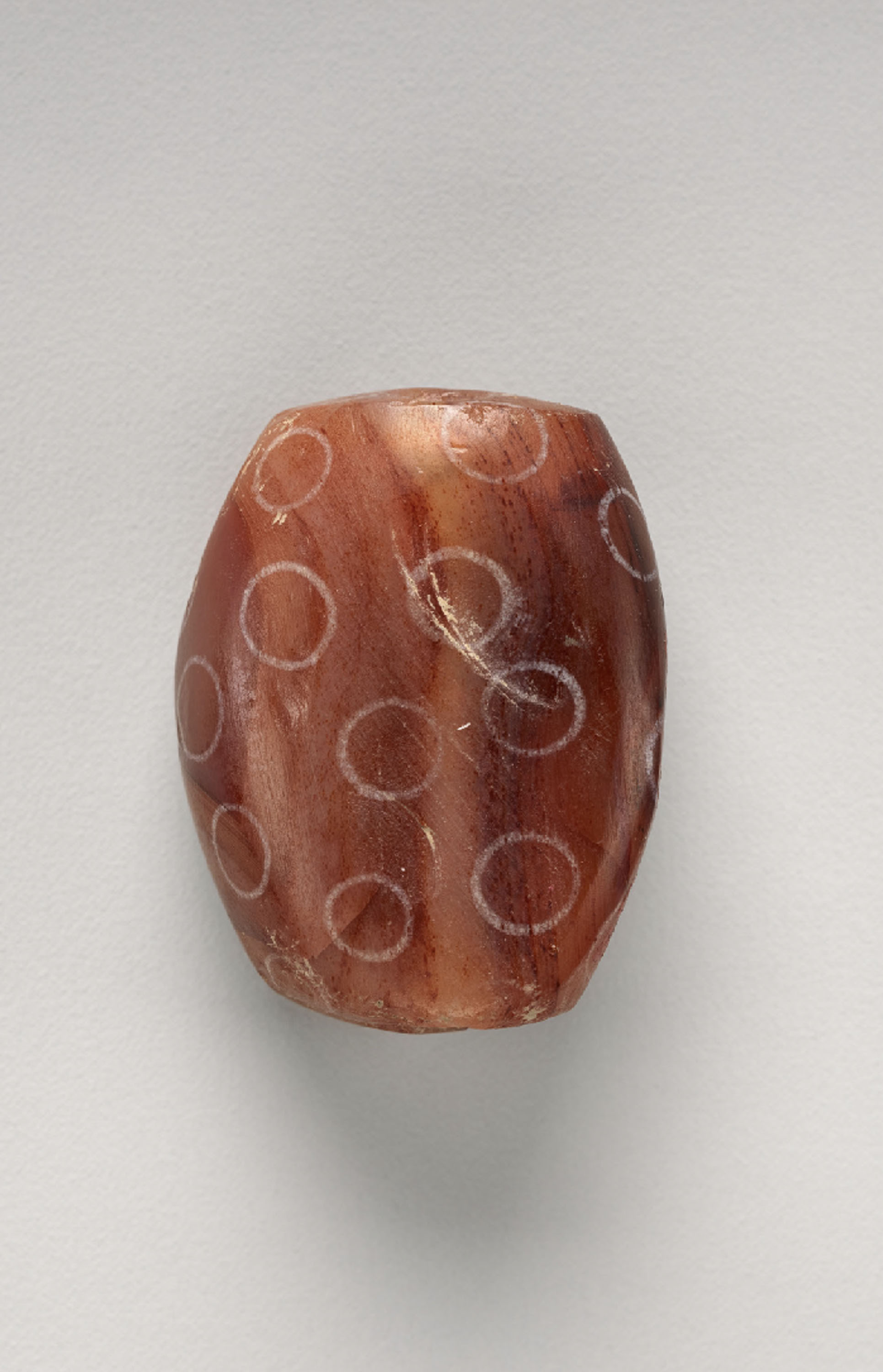 A small reddish-brown carved bead with a smooth surface, decorated with light circular patterns across its sides.