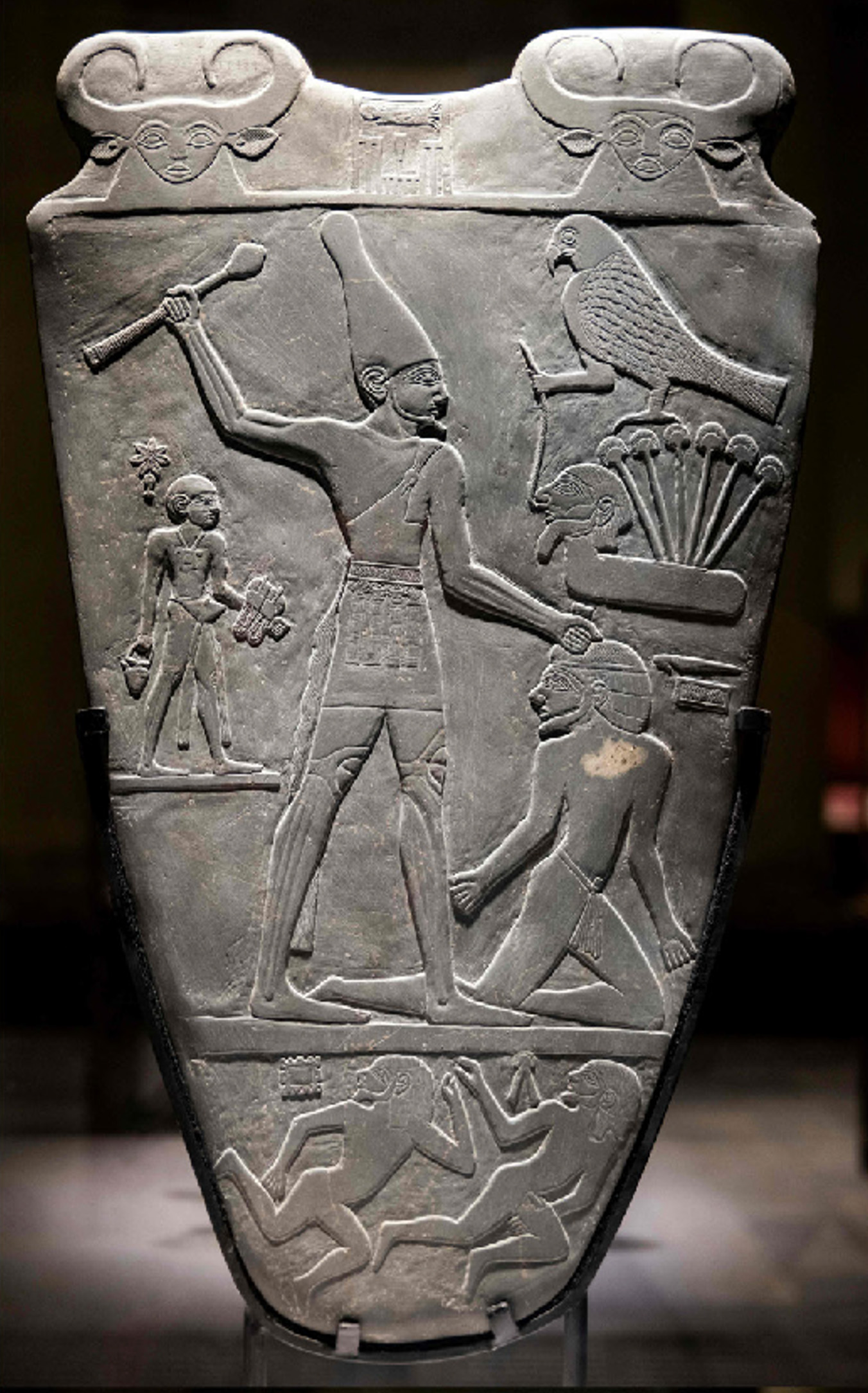 An ancient carved stone palette with detailed relief figures, showing a tall central figure wearing a crown, smaller human figures, animals, and symbolic imagery arranged around them.
