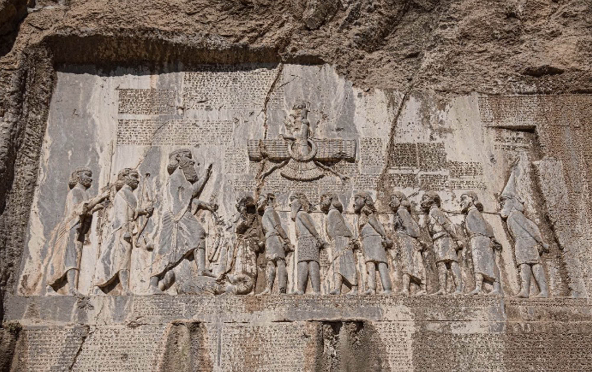 A monumental stone carving in a rock wall of a king before a row of bound prisoners. There is text carved into the wall.