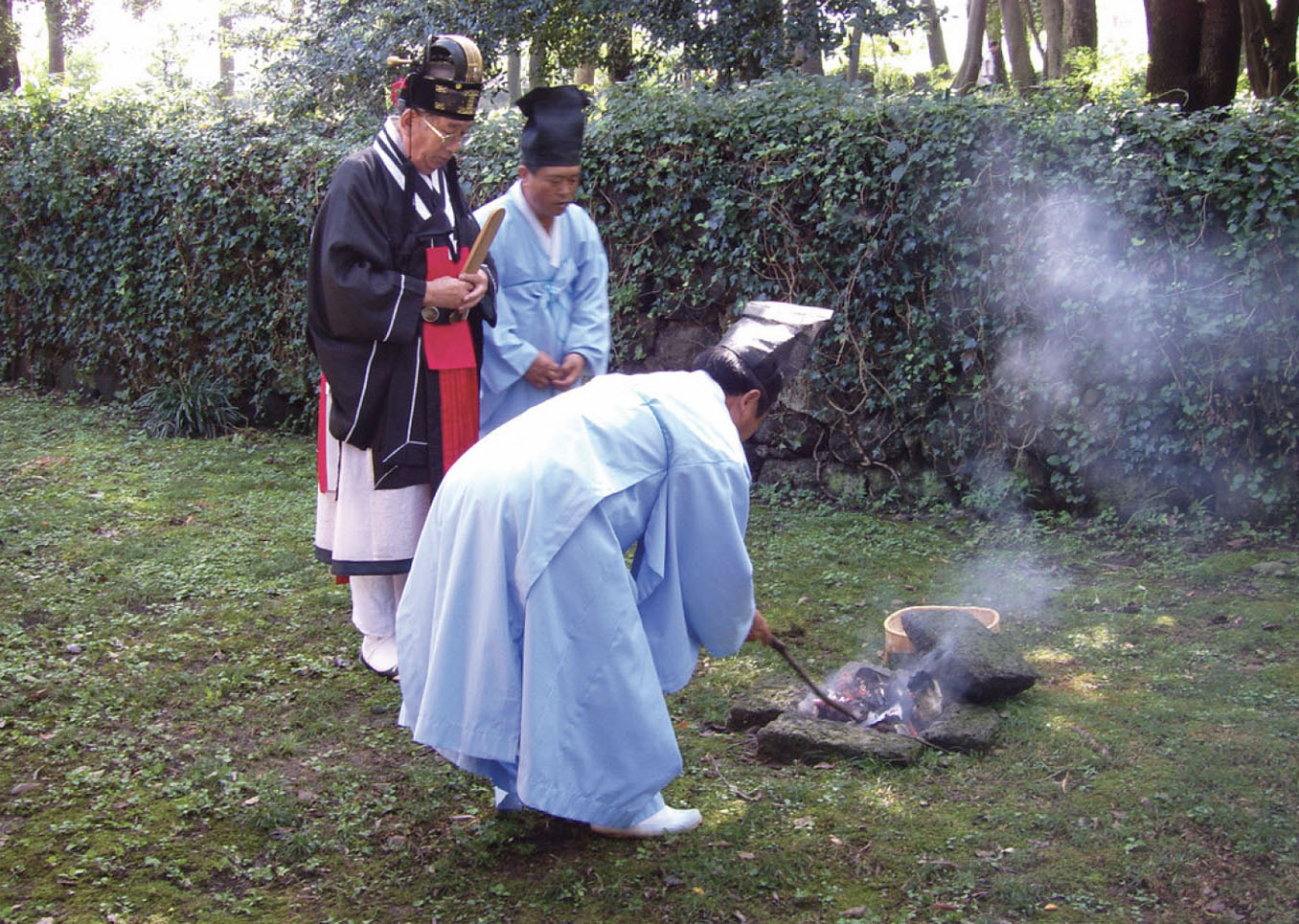 Three men stand around a small fire bit, where tablets are smoldering. One man is poking at the fire pit with a stick