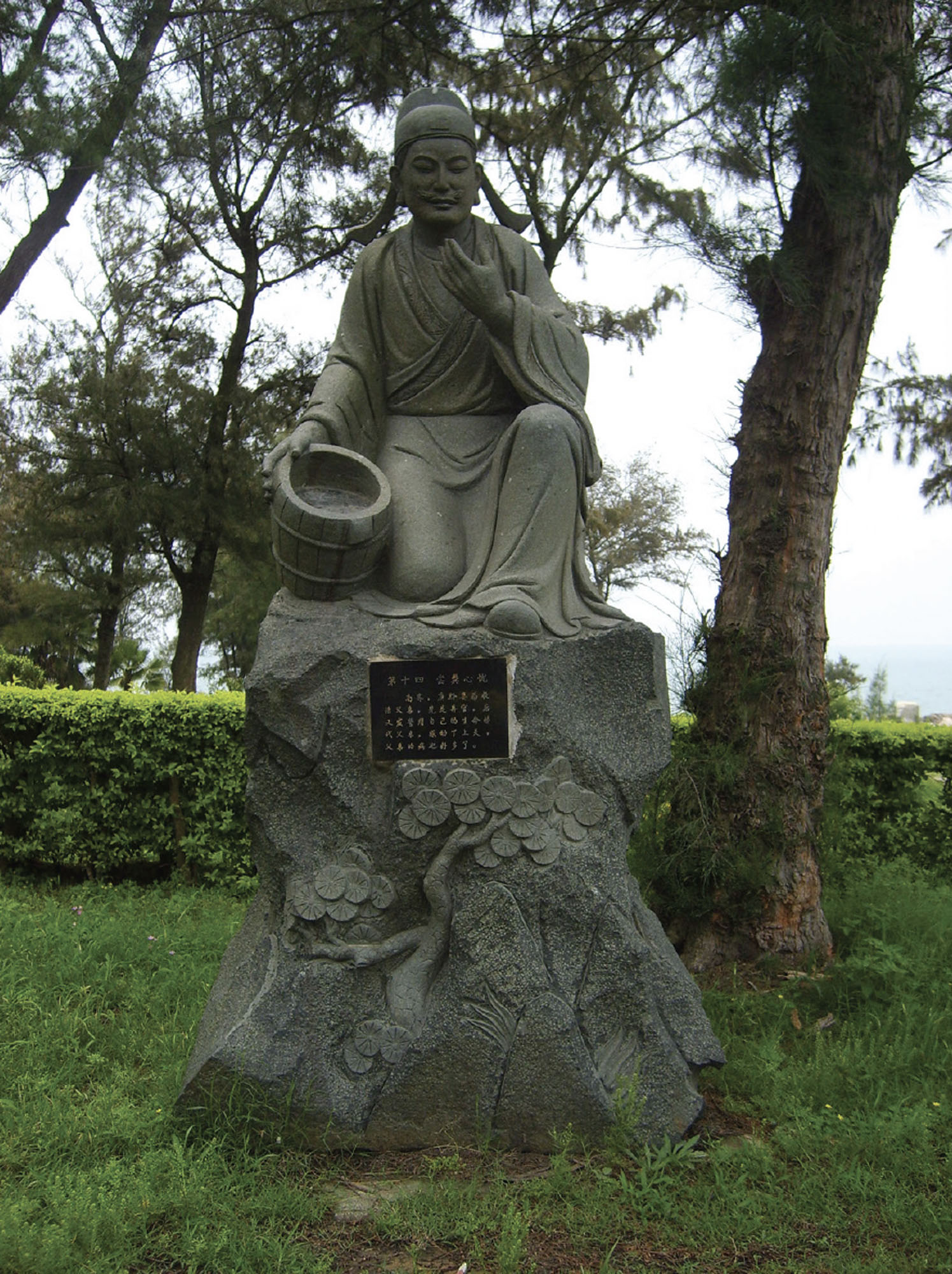 A sculpture of a kneeling person. One hand is raised, and the other is holding a bucket. The sculpture is in a park or garden, surrounded by green grass