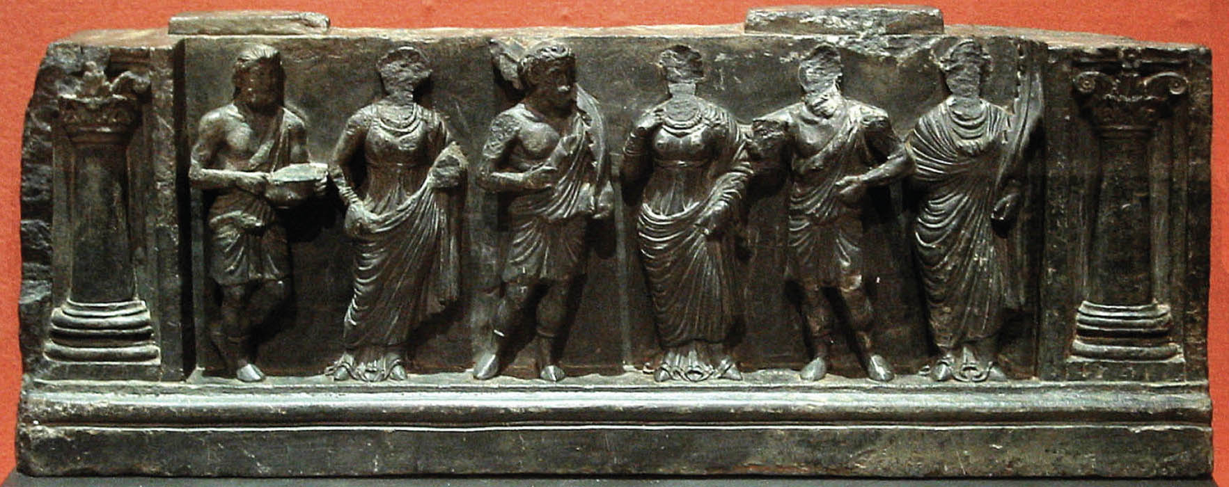 A stone carving of three men and three women, standing in a line, with pillars at either side. Both the men and women are wearing draped clothing. All but two of the people’s faces have fallen off or broken due to age.