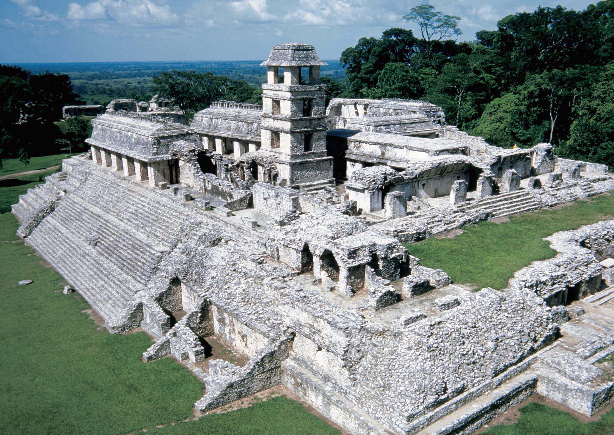 Aerial view of the stone ruins of a Mayan city. The ruins are well preserved, revealing what was a pyramid-like structure with steps leading up to a peristyle. In the middle of the courtyard is a tall, stone building.