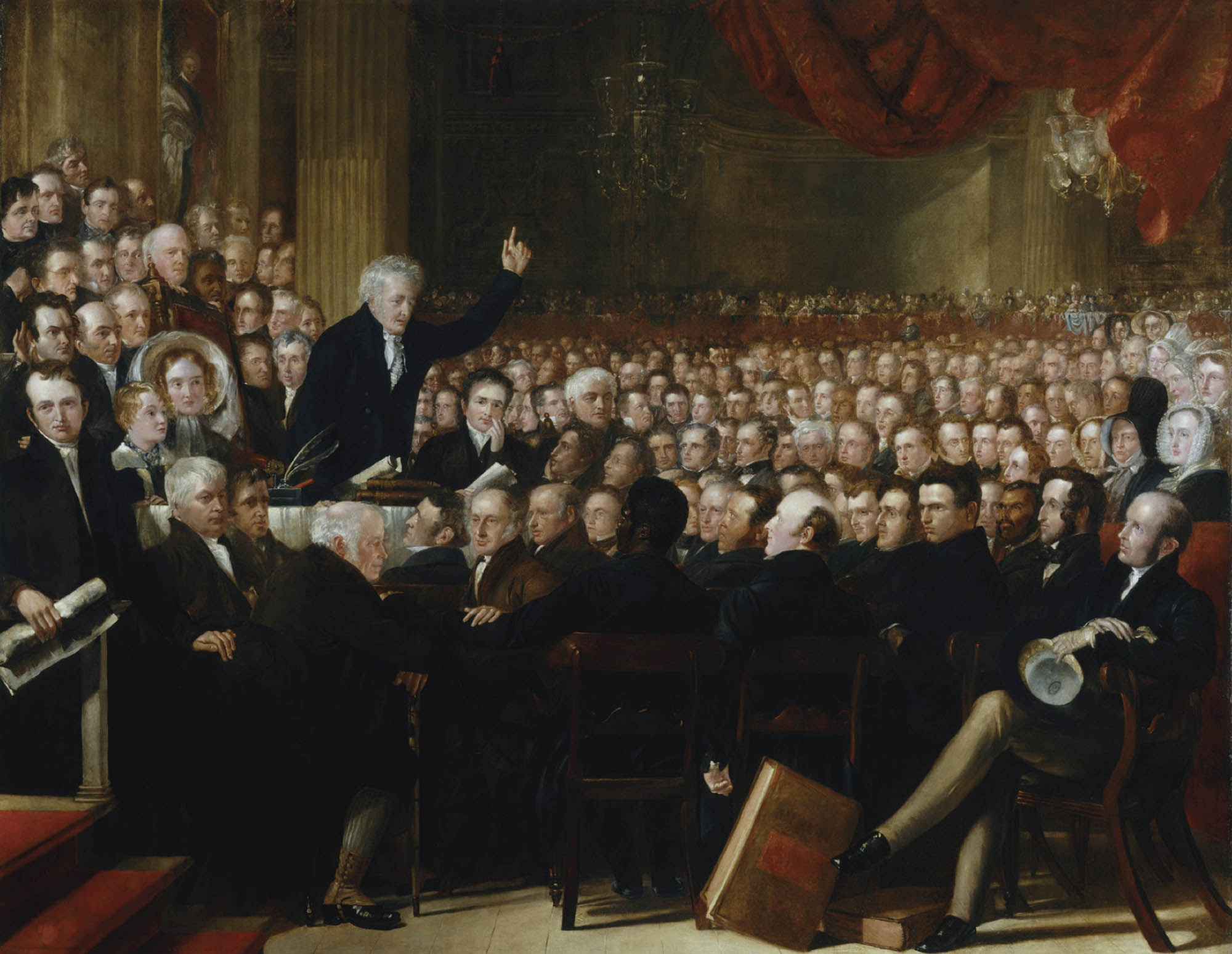 A painting of a large group of people meeting in a grand room. One person is standing, presumably speaking to the room, with one hand raised in the air.