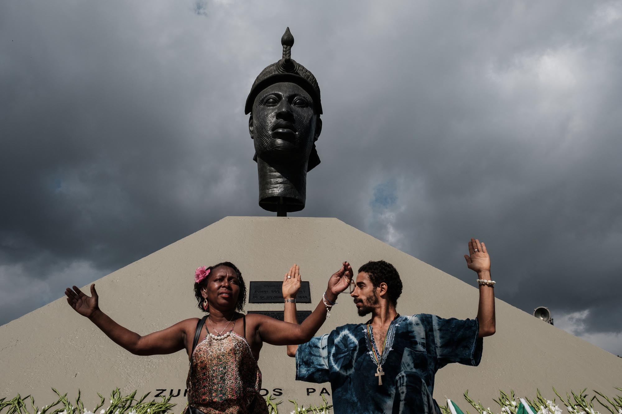 Photograph of two people stand with arms raised in front of a monument, in Rio de Janeiro, dedicated to the slaves killed in battle in 1695.