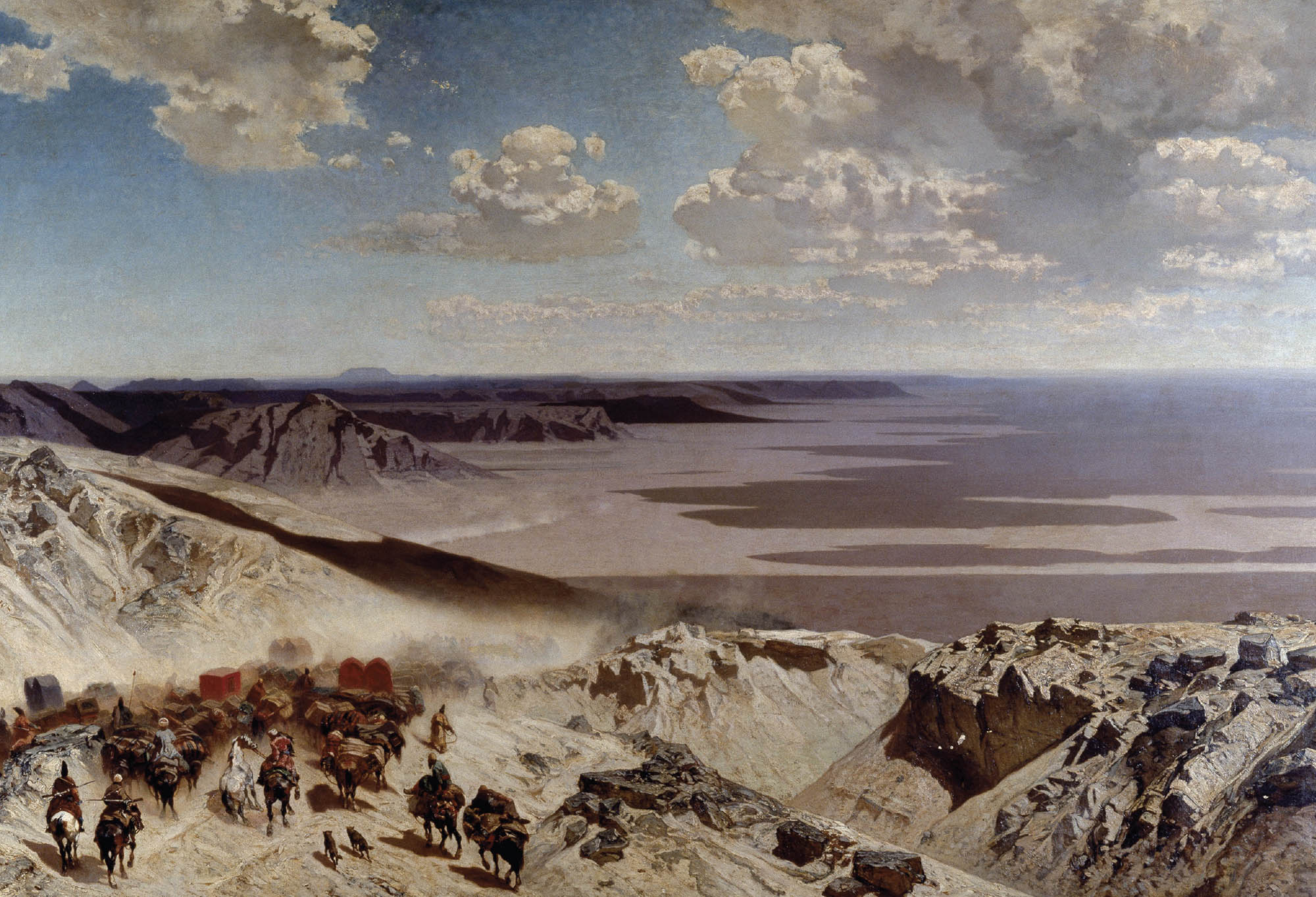 A vast and mountainous desert landscape under blue skies with large white clouds that cast dramatic shadows on the desert below. In the foreground there is a group of people on horses and other animals who have large loads of goods strapped onto them.