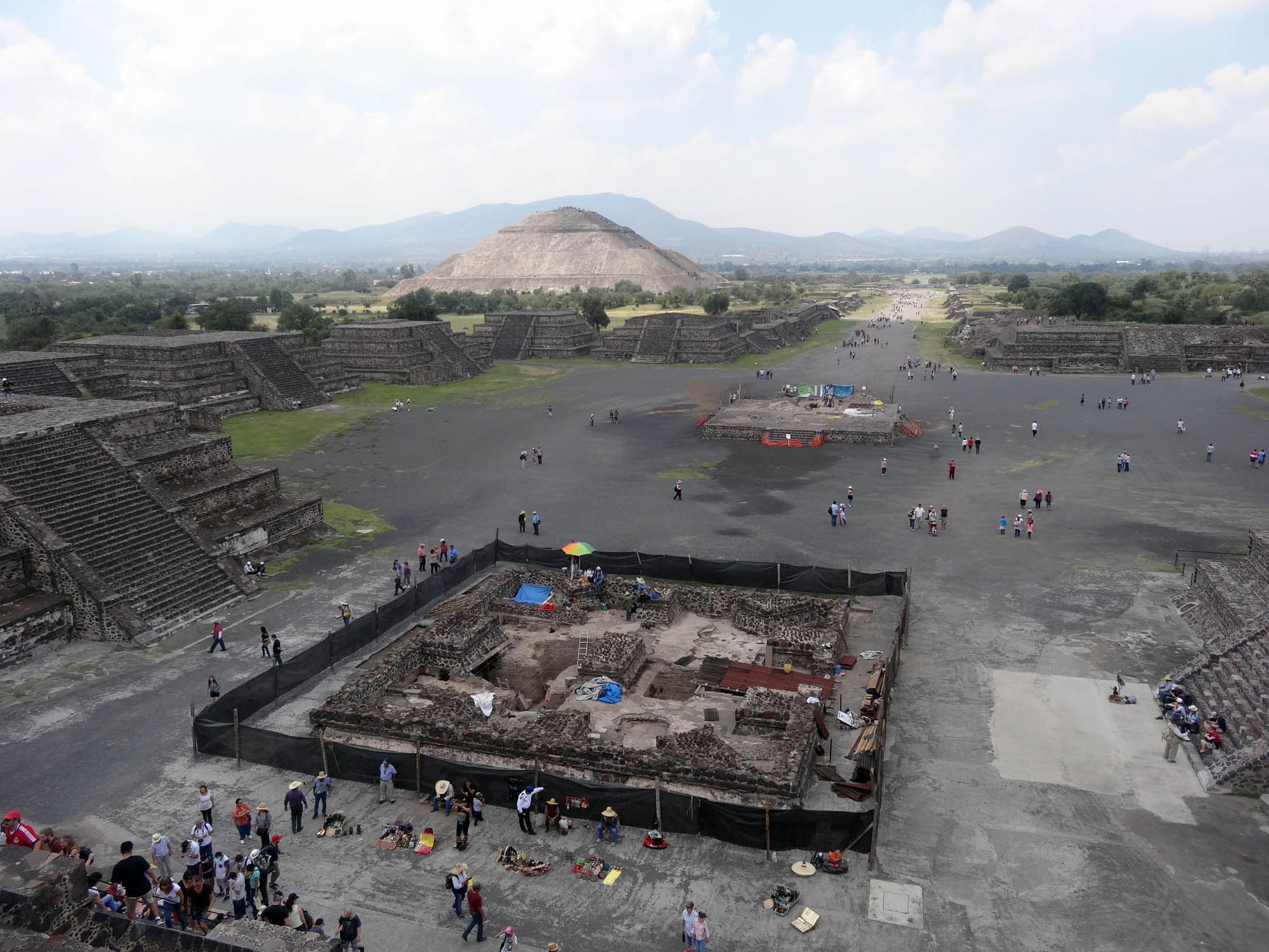 In the center of the city of Teotihuacan, people crowd around an archaeological site. Inside the site we can see remnants of brick structures. The site has a black fence around it.