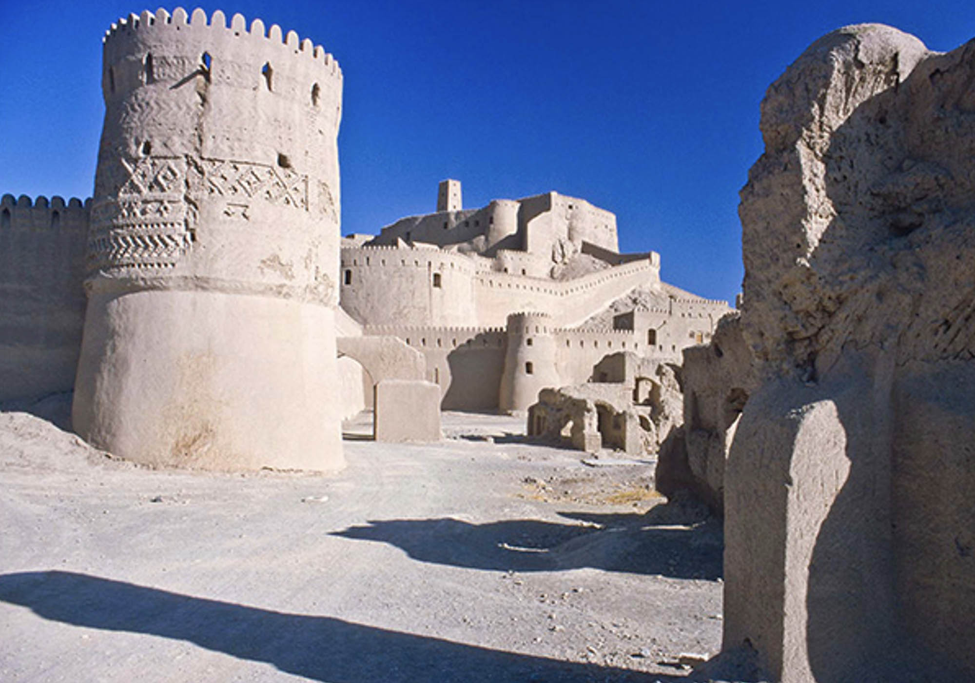 The outer walls of the Safavid Bam citadel, as well as some of the interior buildings, the sky is blue and there are shadows cast by the buildings.