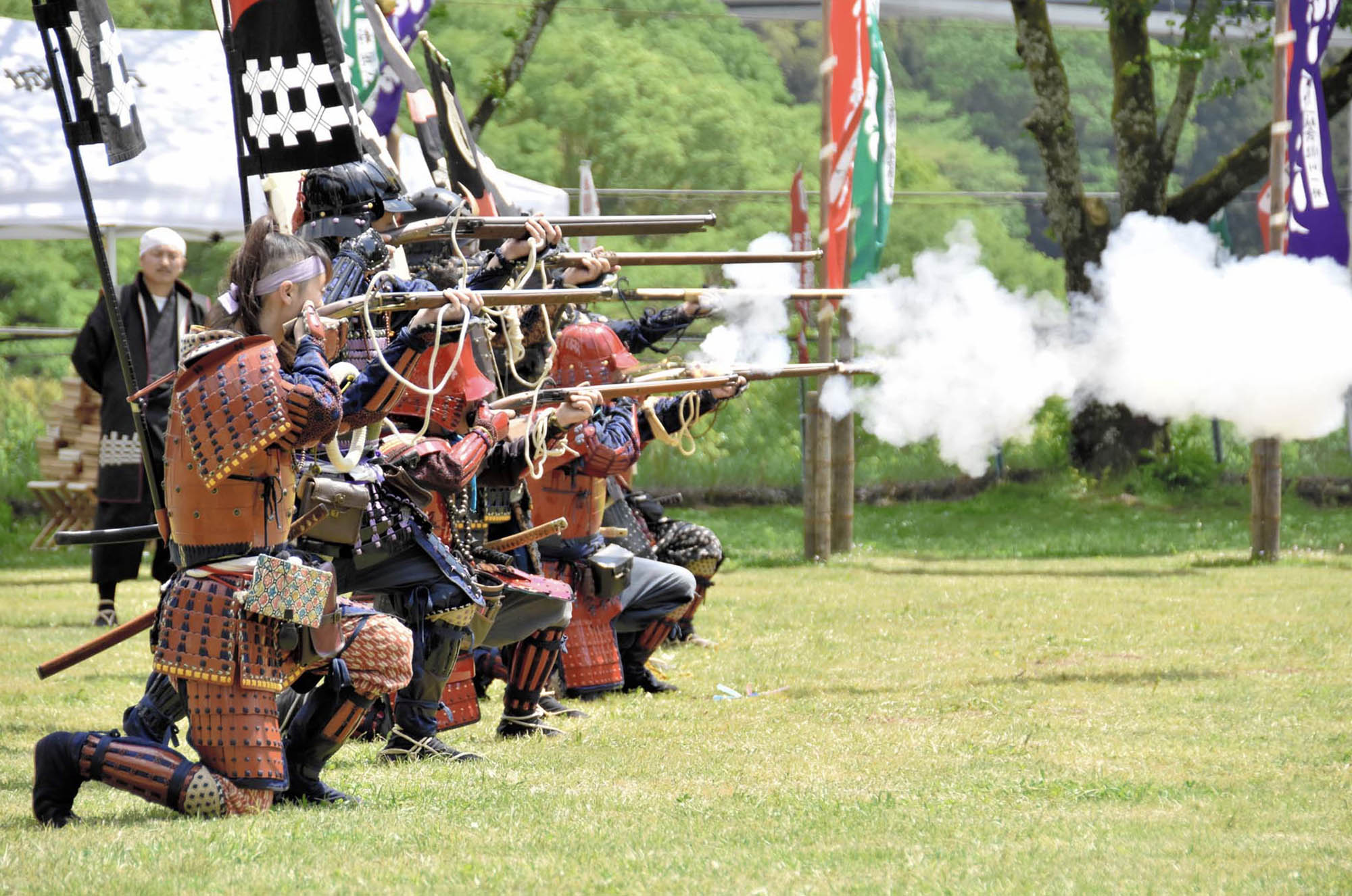A group of people in Japanese armor kneeling in a grassy field while firing matchlock rifles.