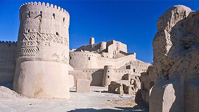 The outer walls of the Safavid Bam citadel, as well as some of the interior buildings, the sky is blue and there are shadows cast by the buildings.