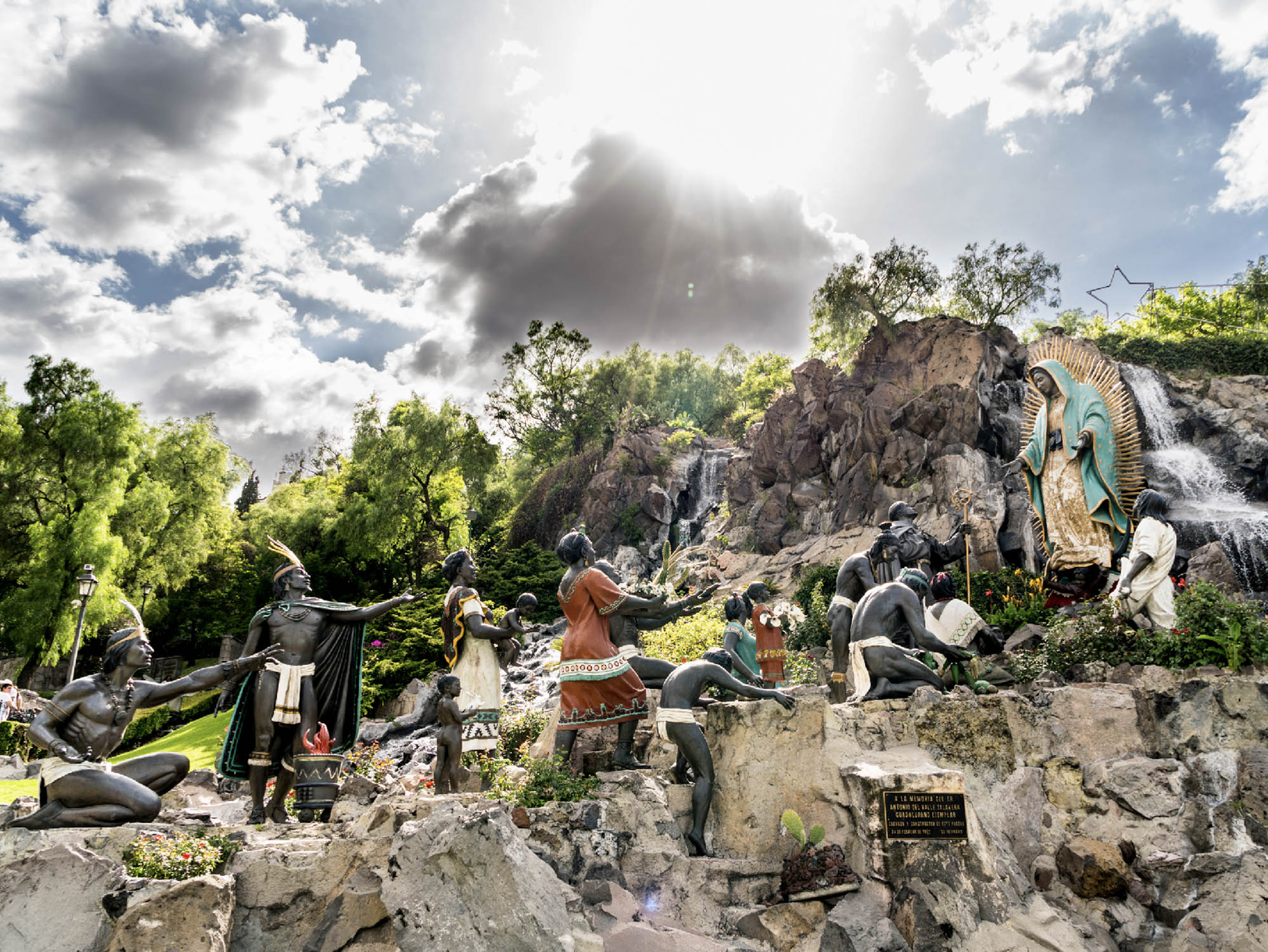 A group of statues on the Tepeyac Hill in Mexico City, the statues are positioned on a rocky terrain with a waterfall amongst a cloudy sky.
